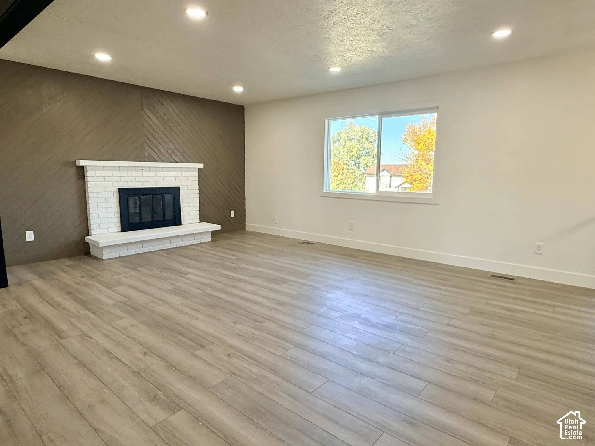 Wood-finish flooring throughout, white-painted brick fireplace with a raised hearth, diagonal wood-plank accent wall, recessed lighting, and large sliding window