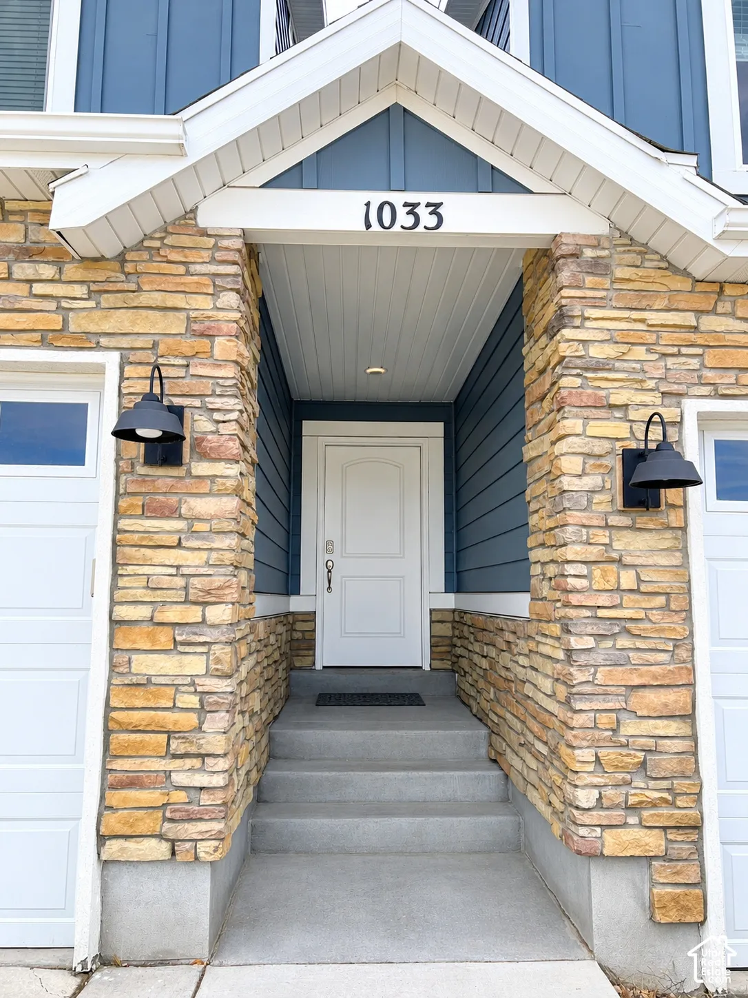 Exterior facade featuring stacked stone masonry, blue horizontal siding, a gabled roofline, concrete steps, and two wall-mounted metal exterior light fixtures