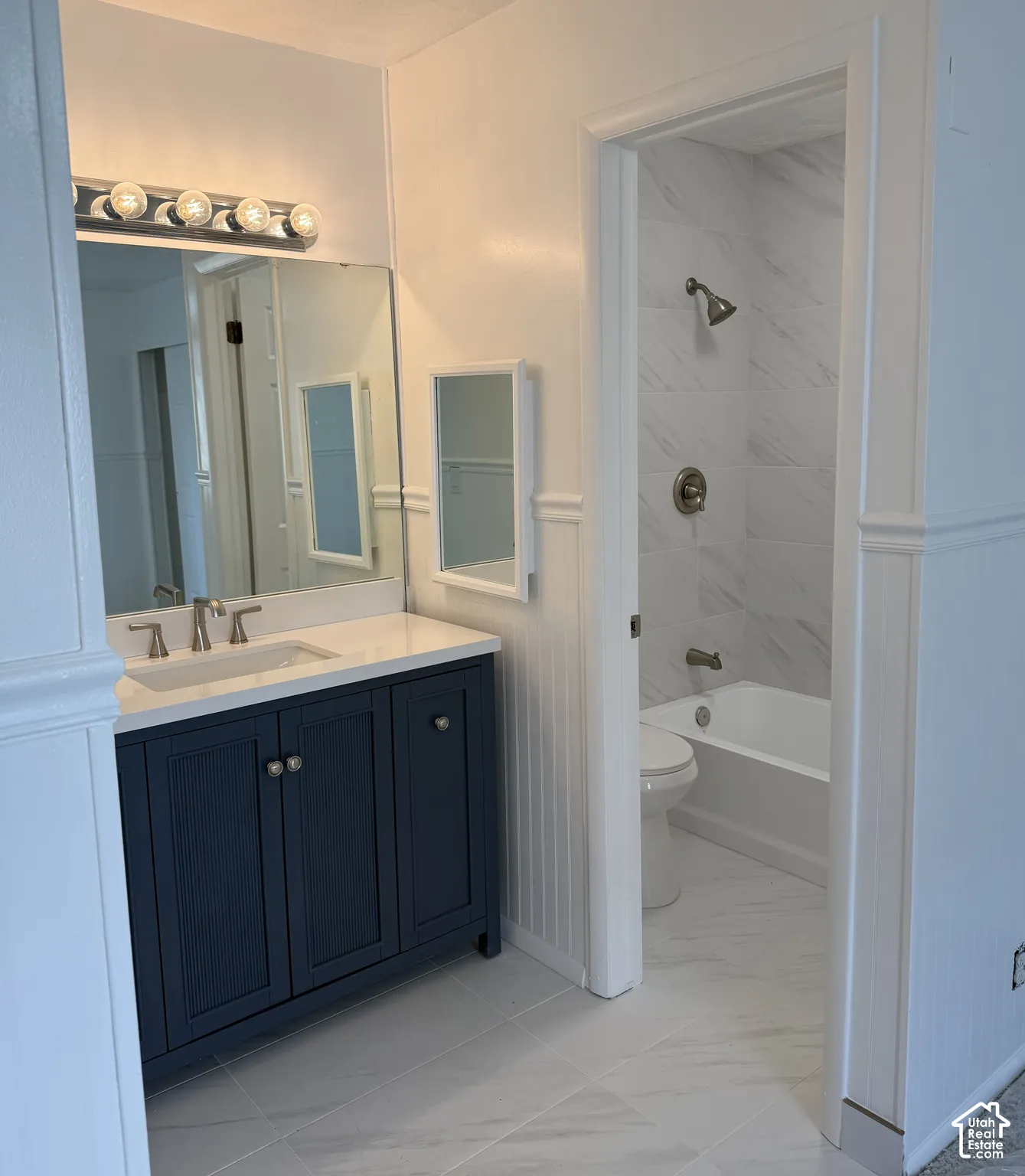 Bathroom featuring a vanity with white countertop, dark cabinetry, wall-to-wall mirror, beadboard wainscoting, and a tiled tub surround