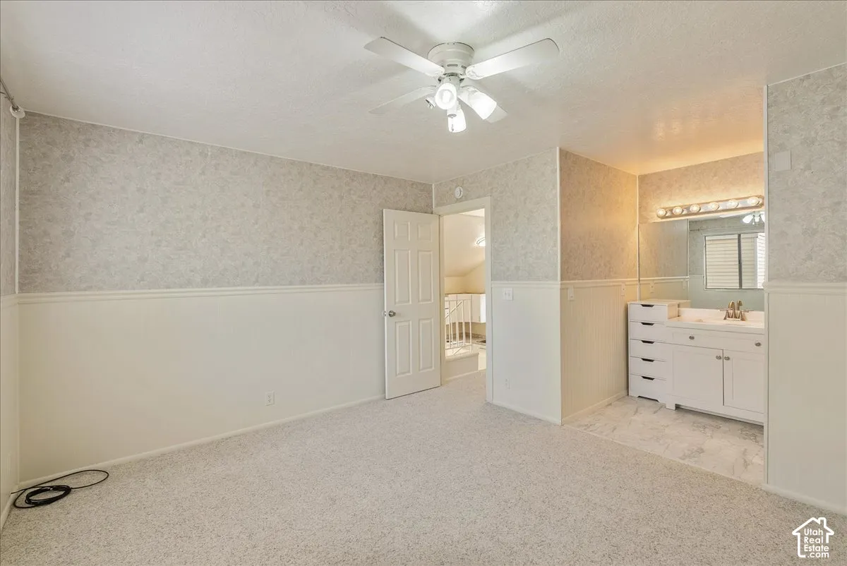 Neutral-toned room featuring patterned wallpaper, chair rail molding, carpeted flooring, a ceiling fan with integrated lighting, and an vanity area with a wall-to-wall mirror