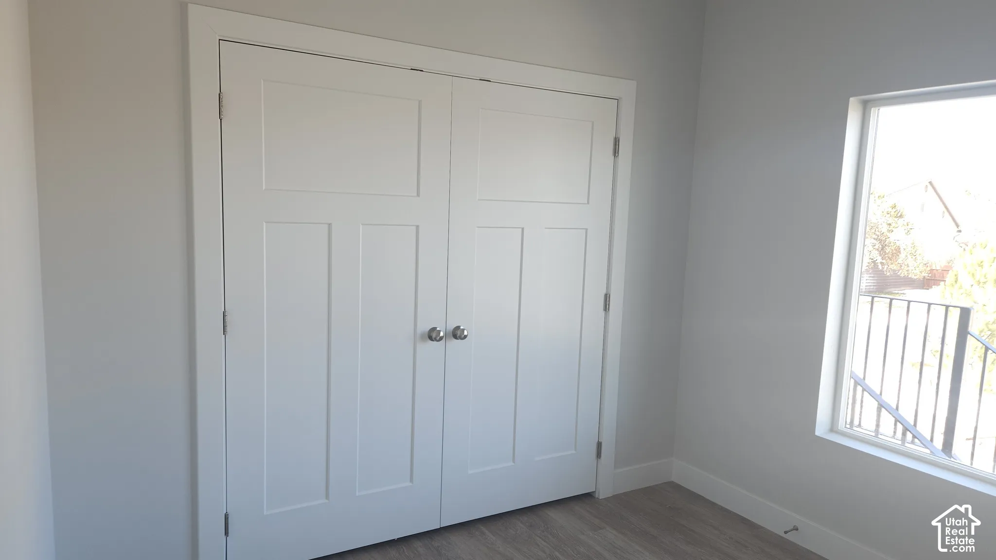 Double-door closet with recessed shaker-style panels and chrome hardware, featuring neutral wall finishes and wood-finish flooring with a window providing natural lighting