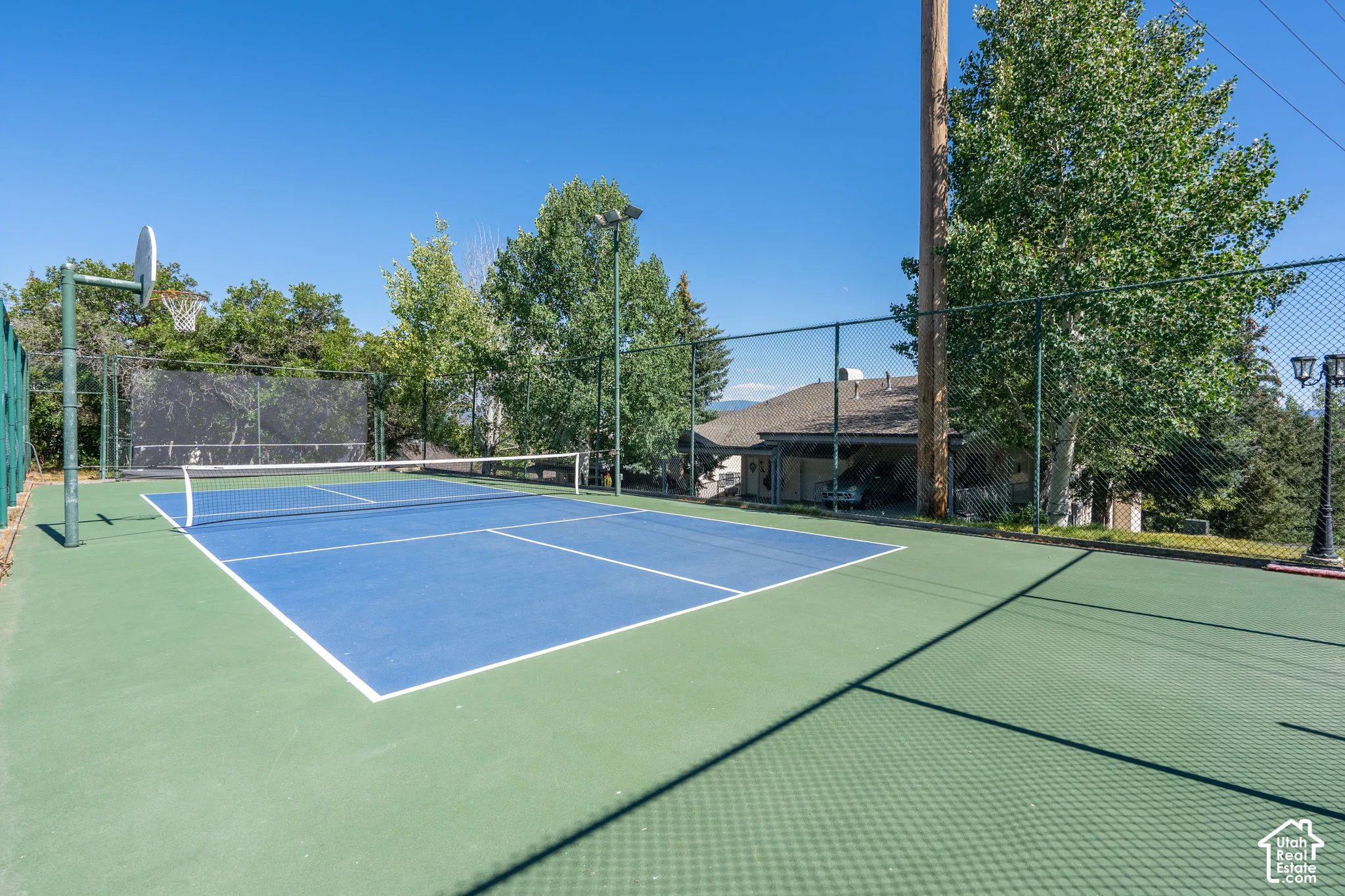 Sport court featuring a multi-use blue and green surface, chain-link fencing, overhead pole lighting, and an integrated basketball hoop