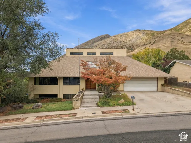 Multi-level exterior featuring stucco siding, a shingled roof, a double-car garage, and concrete walkway, set against a prominent mountain backdrop
