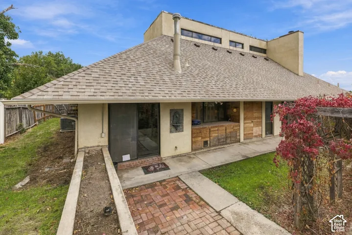 Stucco exterior featuring a tiered roofline, integrated chimney, brick paved walkway, wood-paneled accents, and climbing foliage