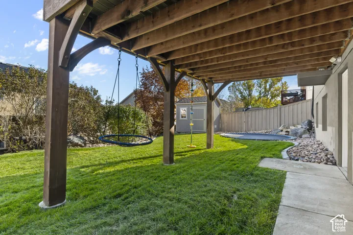 Covered concrete patio featuring timber framing, landscaped lawn, stone border accents, and an attached storage shed