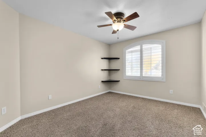 Neutral-toned room featuring plush wall-to-wall carpeting, an arched window with fitted plantation shutters, three built-in corner shelves, and a ceiling fan with integrated lighting