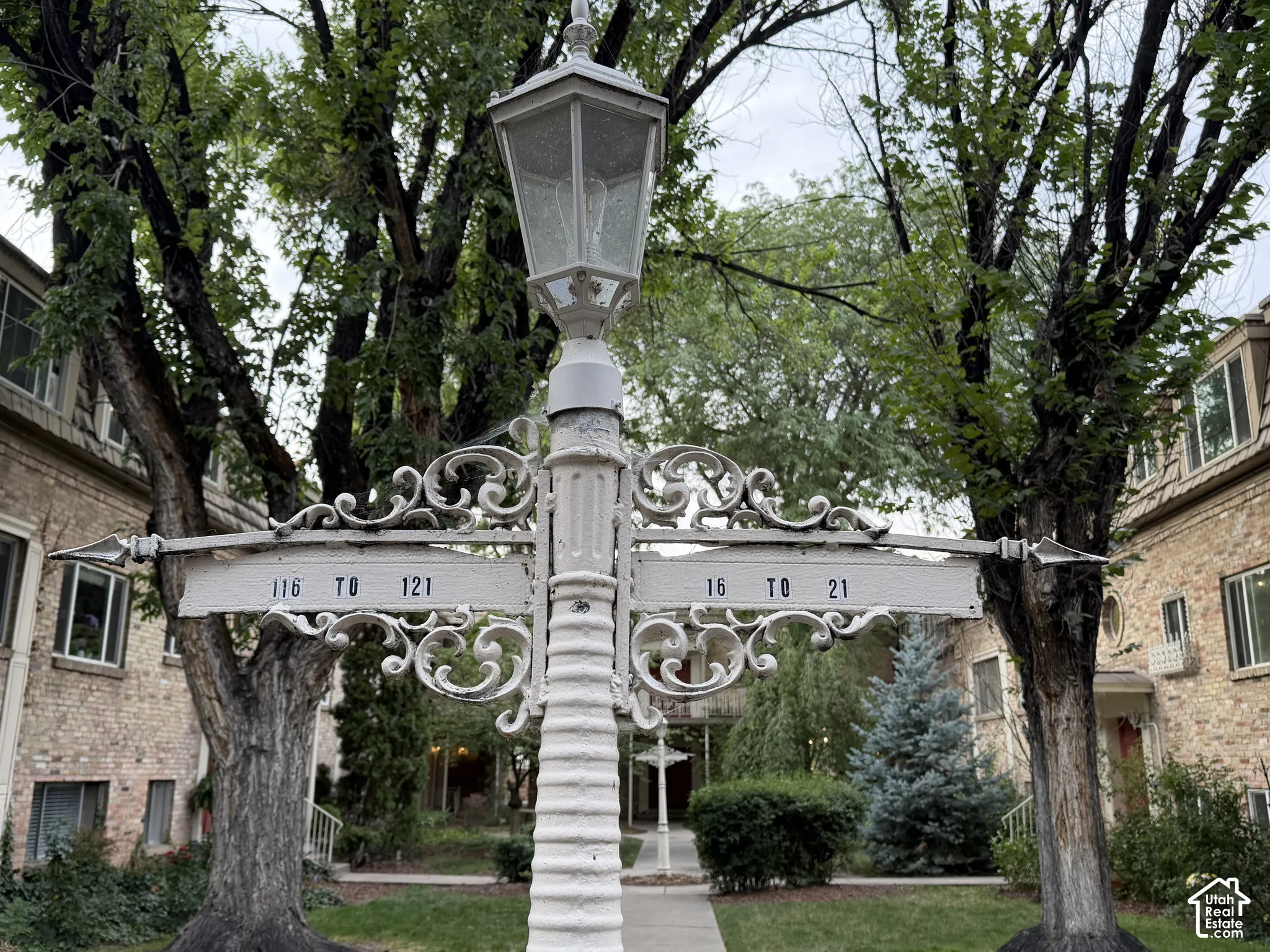 Units are indicated by decorative white metal lamp posts situated amidst mature deciduous trees in front of each building.