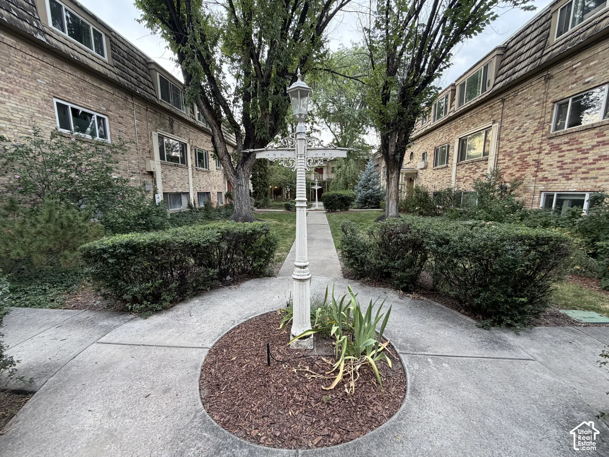 Community central courtyard with mature trees, manicured hedges, a concrete walkway, and the directional lamppost