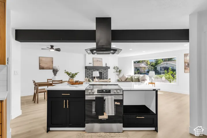 Kitchen island featuring black cabinetry, gold hardware, and a white countertop with an integrated stainless steel range, accented by a black suspended range hood and wood-finish flooring