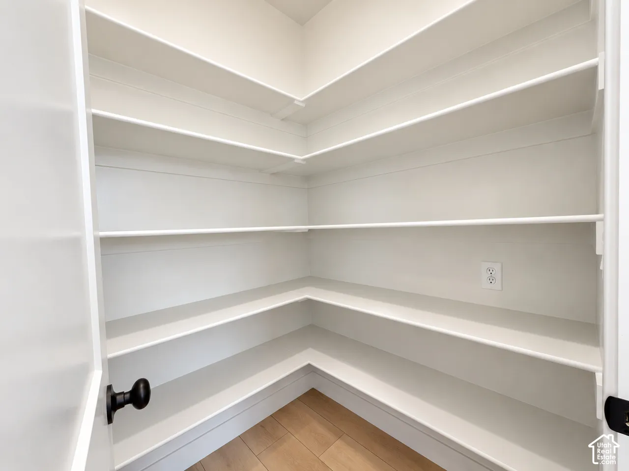 Walk-in storage space featuring wrap-around white shelving, wood-finish flooring, and a wall-mounted electrical outlet