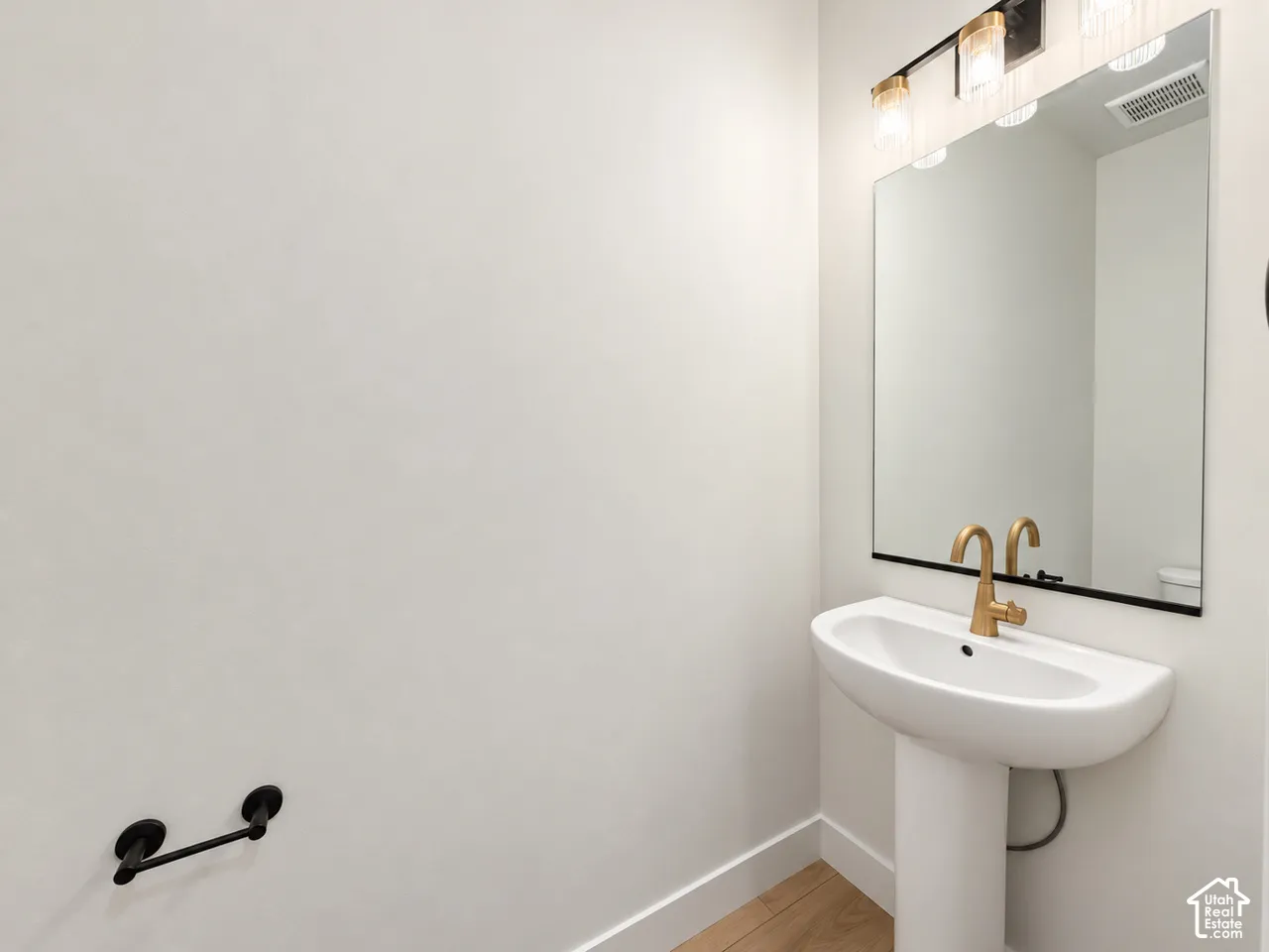 Pedestal sink with a brass-finish faucet, wall-mounted matte black hardware, large rectangular mirror, and three-light brass vanity fixture