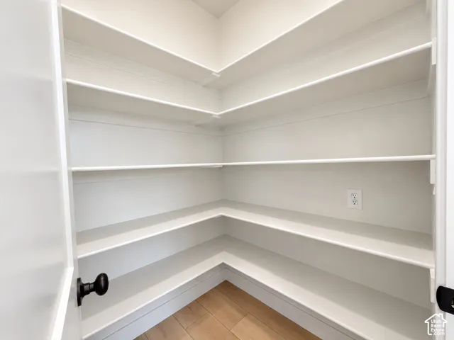 Walk-in storage space featuring wrap-around white shelving, wood-finish flooring, and a wall-mounted electrical outlet