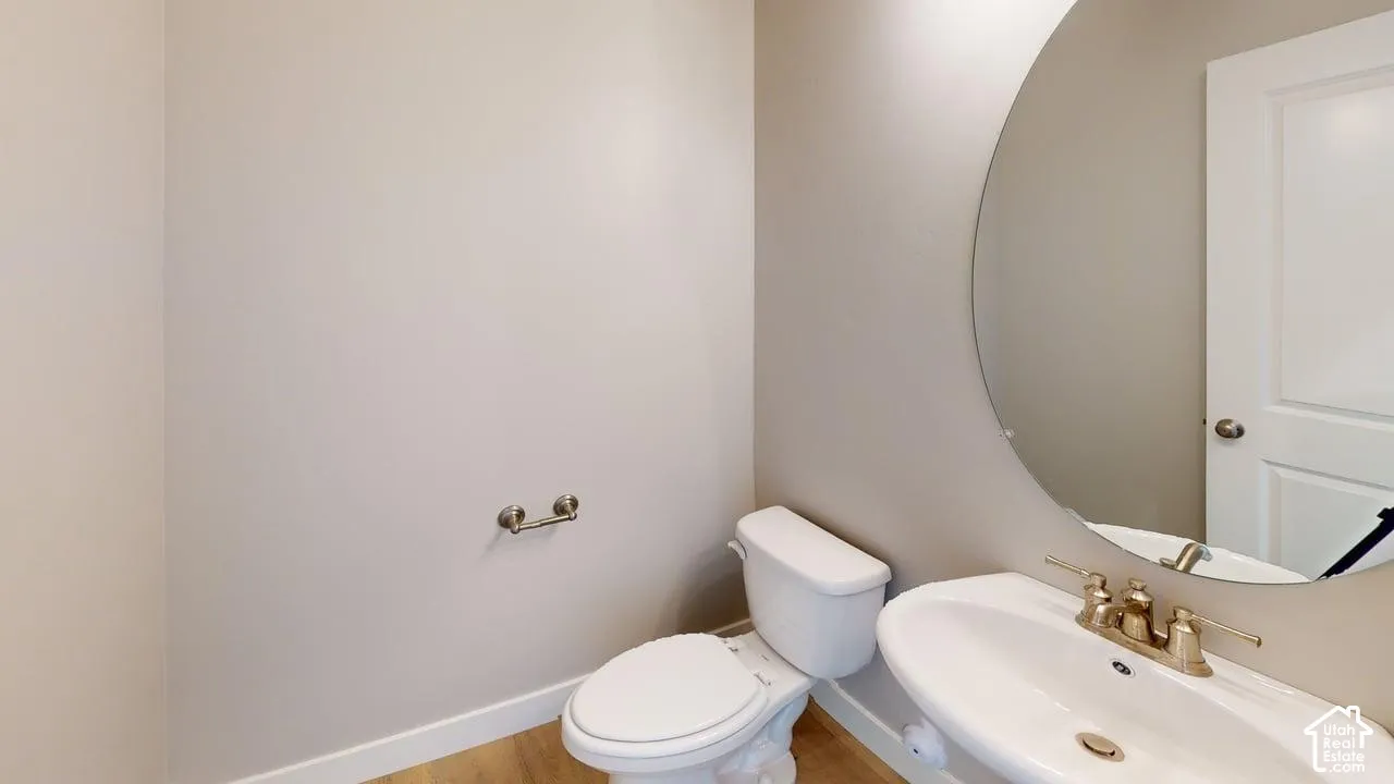 Wall-mounted circular mirror, pedestal sink with brass-finish faucet, elongated toilet, and wood-finish flooring