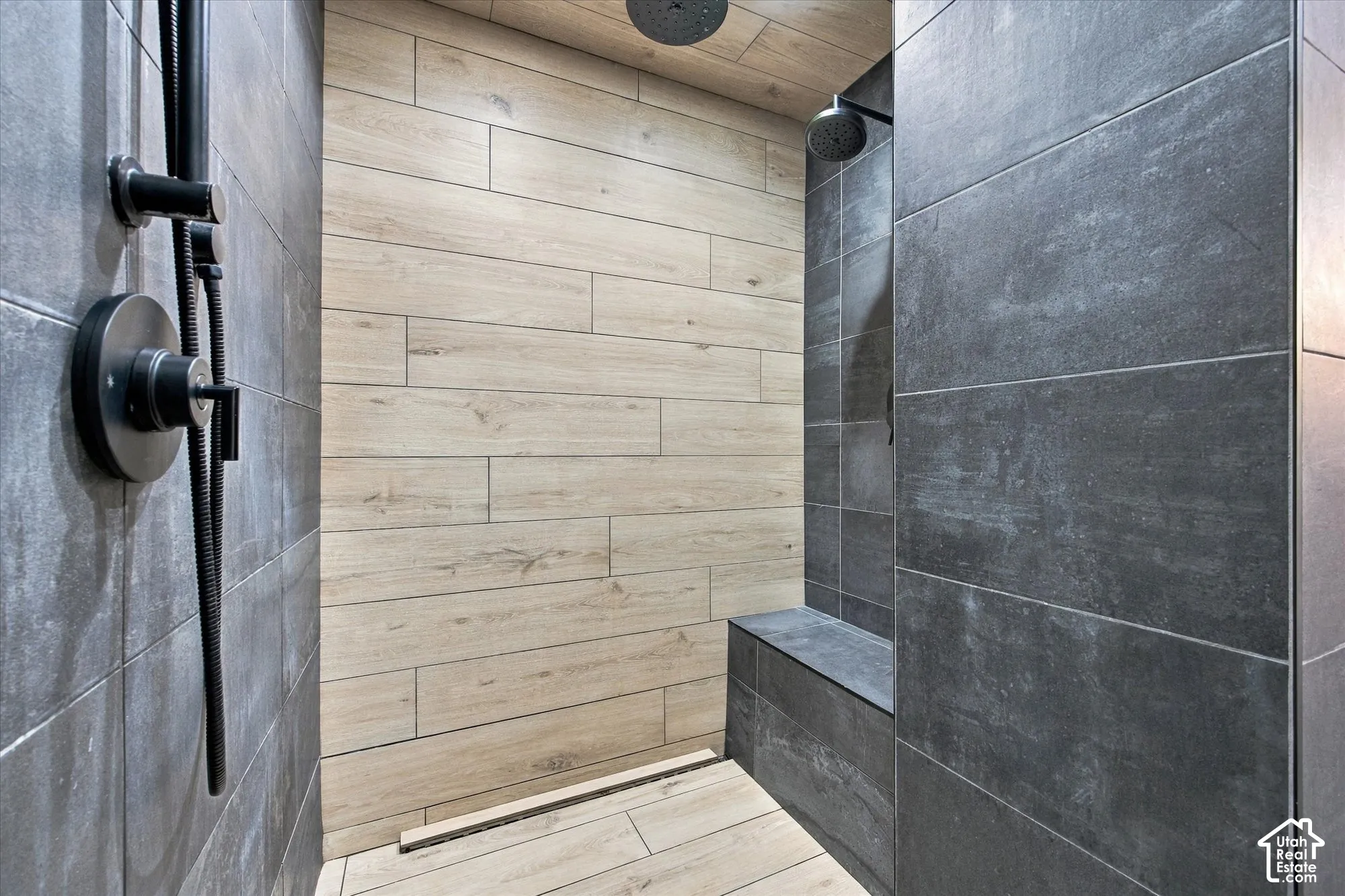 Walk-in shower featuring matte black fixtures, dual overhead rainfall showerheads, wood-finish tile accent wall, large-format grey tile, and a built-in stone-finish bench