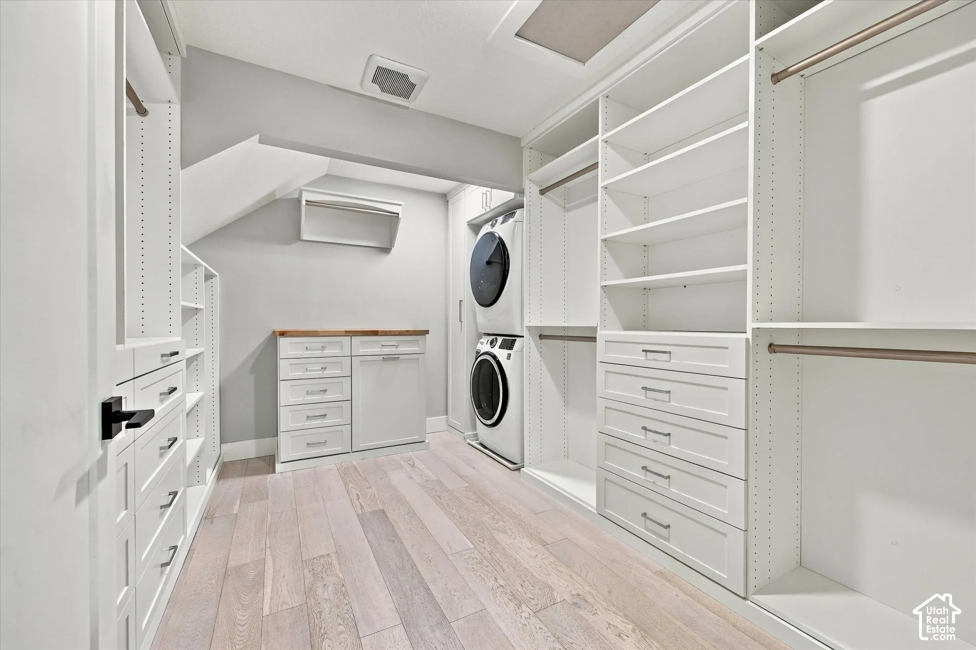 Walk-in dressing area featuring extensive custom built-in white cabinetry, integrated drawers, wood-finish flooring, and a dedicated laundry alcove with a stacked washer and dryer