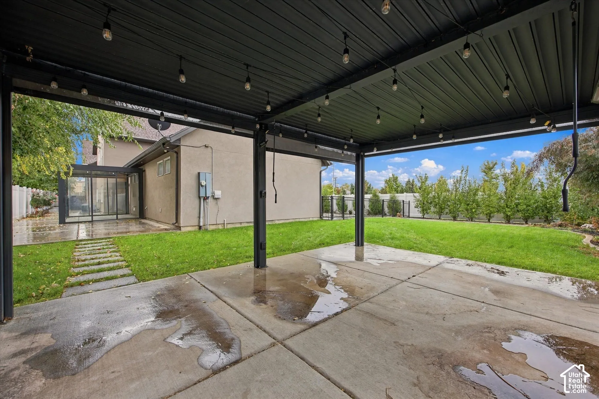 Covered outdoor patio with a black metal pergola, string lighting, concrete flooring, and an expansive lawn