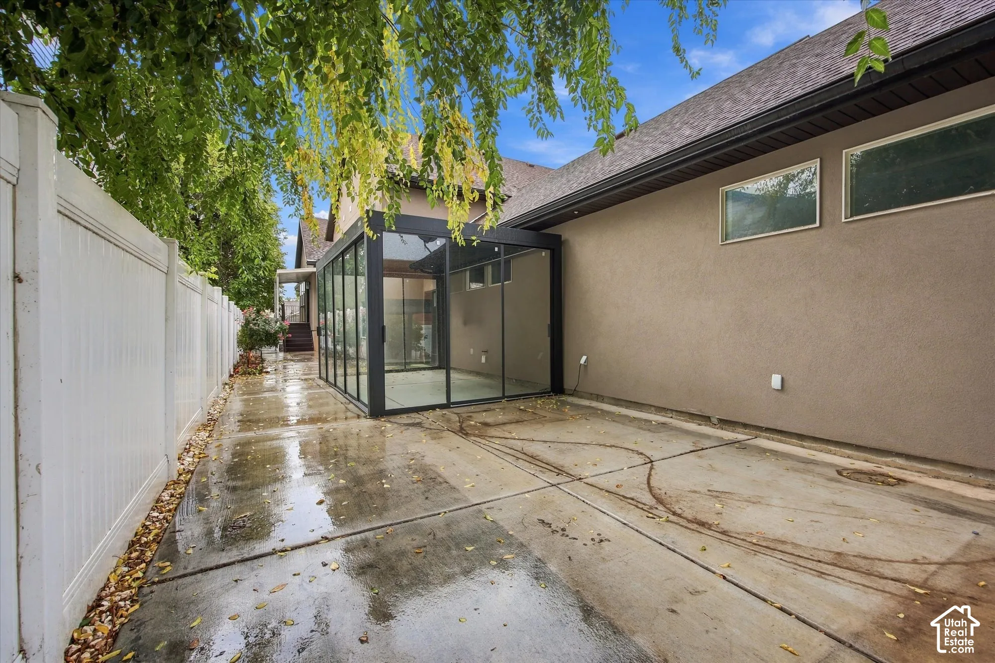 Enclosed glass sunroom featuring a dark metal frame, set upon a concrete patio surface, bordered by a white vinyl privacy fence and stucco exterior walls