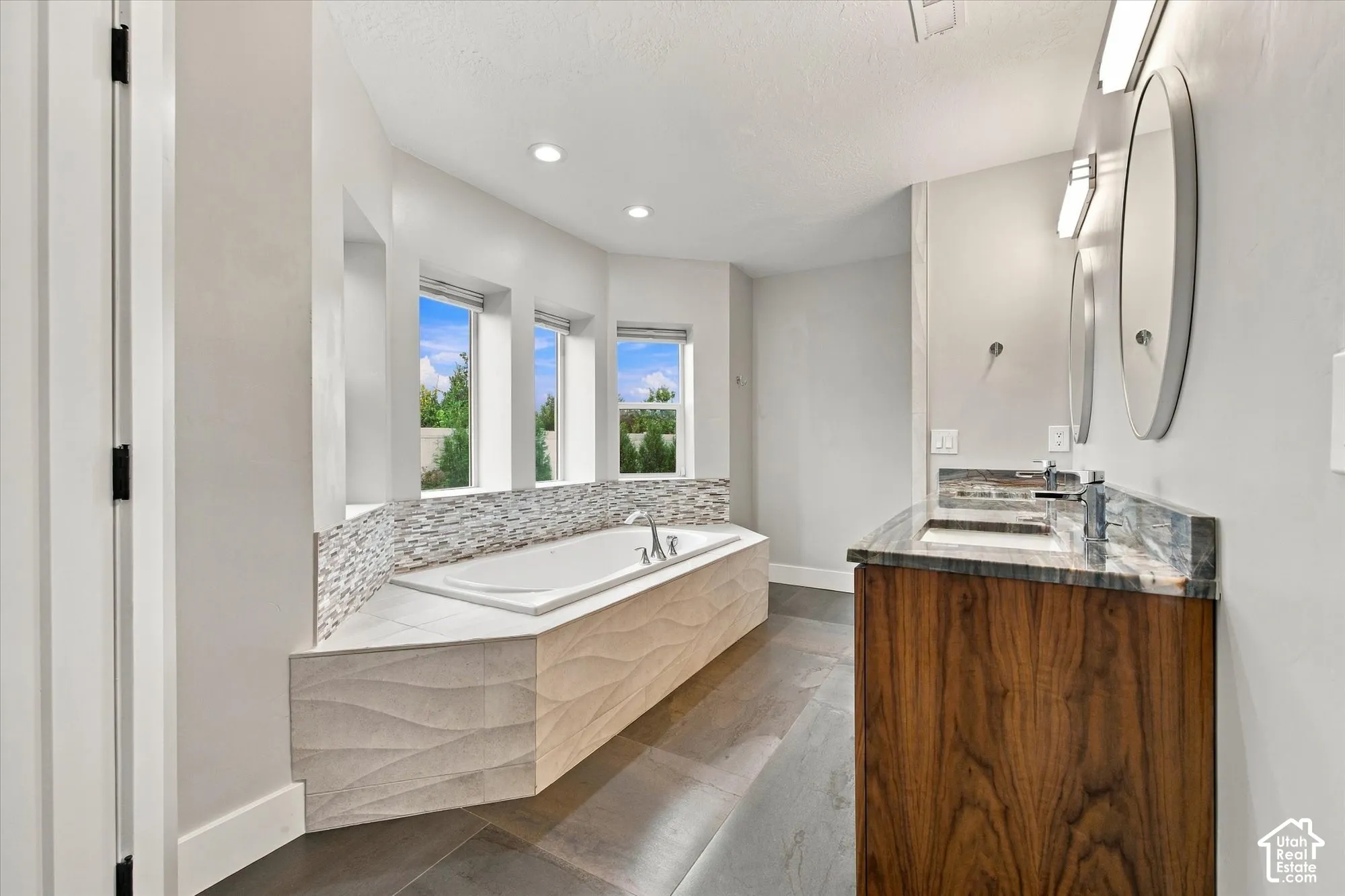 Built-in soaking tub with a textured tile surround, dual-sink vanity with stone-finish countertops, wood-finish cabinetry, and recessed lighting