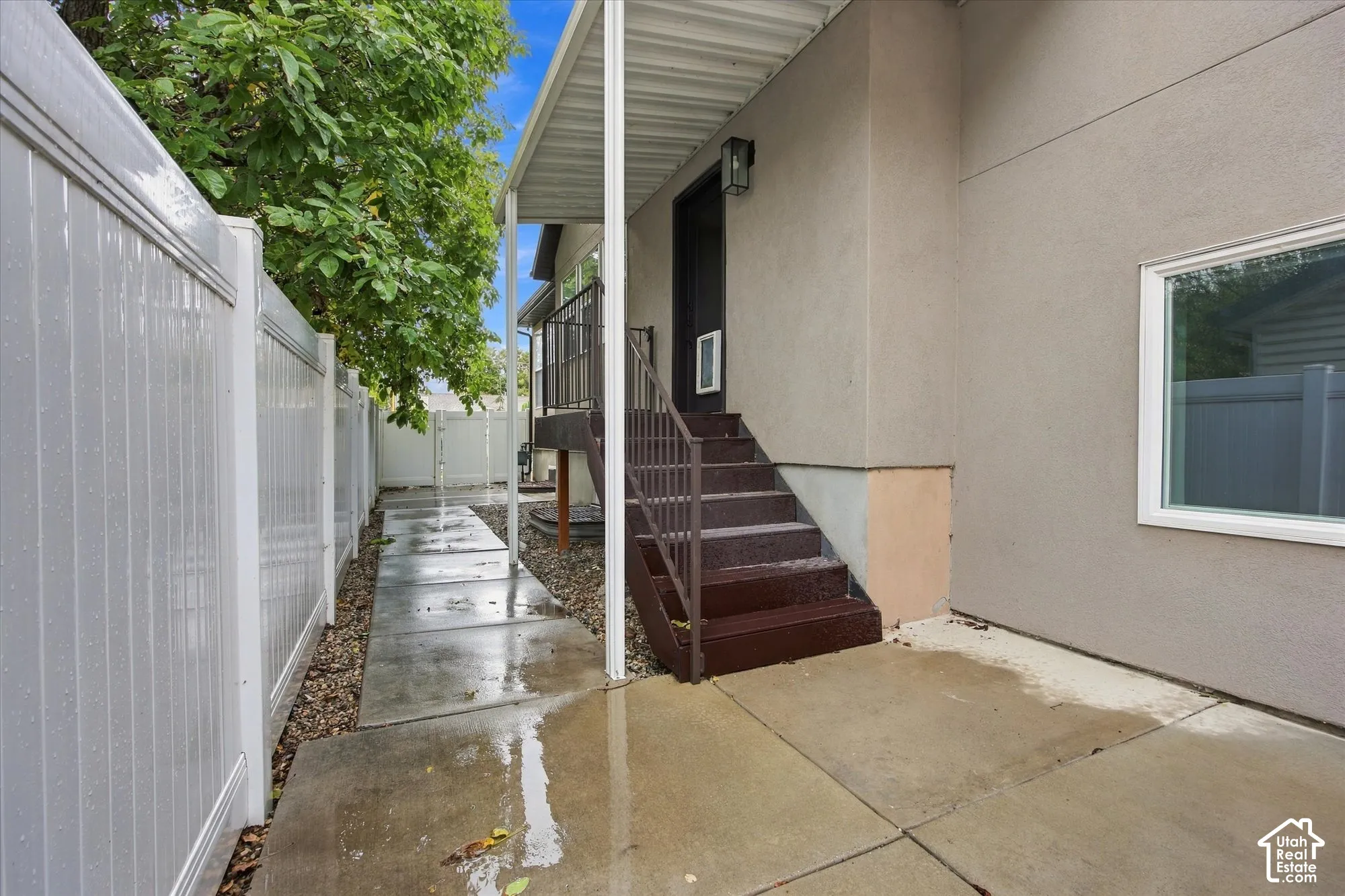 Side exterior featuring a concrete patio, white vinyl privacy fencing, covered entry steps with metal handrails, and low-maintenance stone landscaping