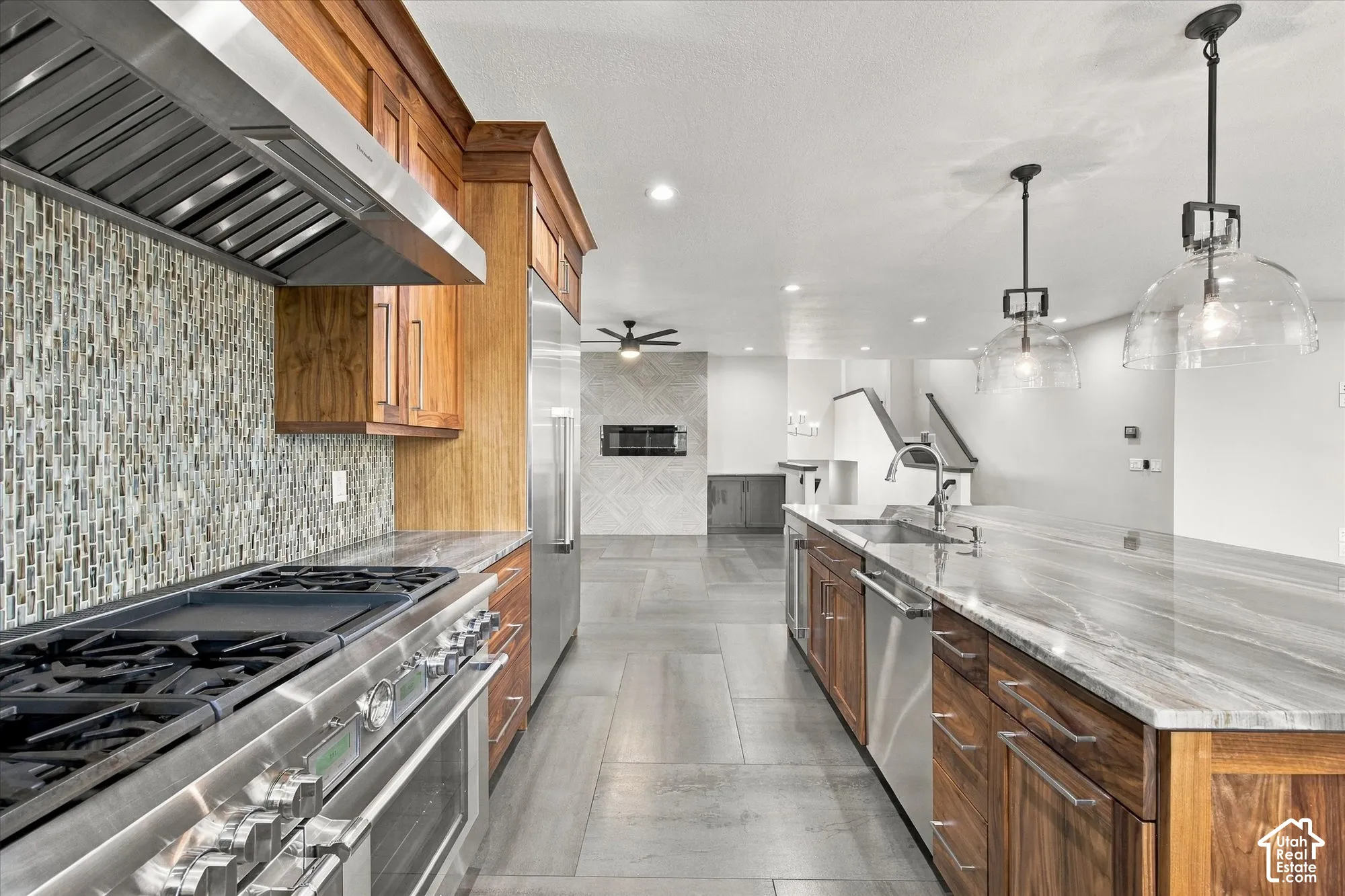 Kitchen featuring wood cabinetry, natural stone countertops, stainless steel appliances, multi-toned tile backsplash, and oversized pendant lighting