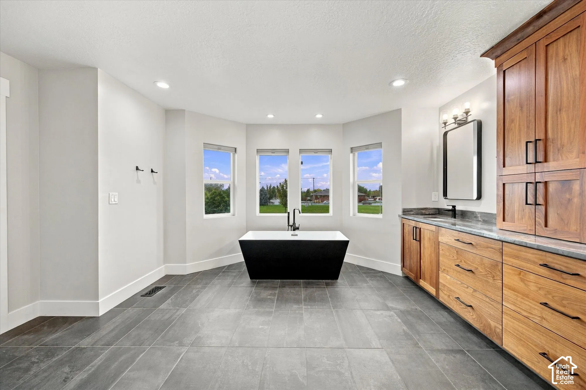 Freestanding black soaking tub, dual-sink vanity with natural wood cabinetry, stone-finish countertops, recessed lighting, and large-format tile flooring