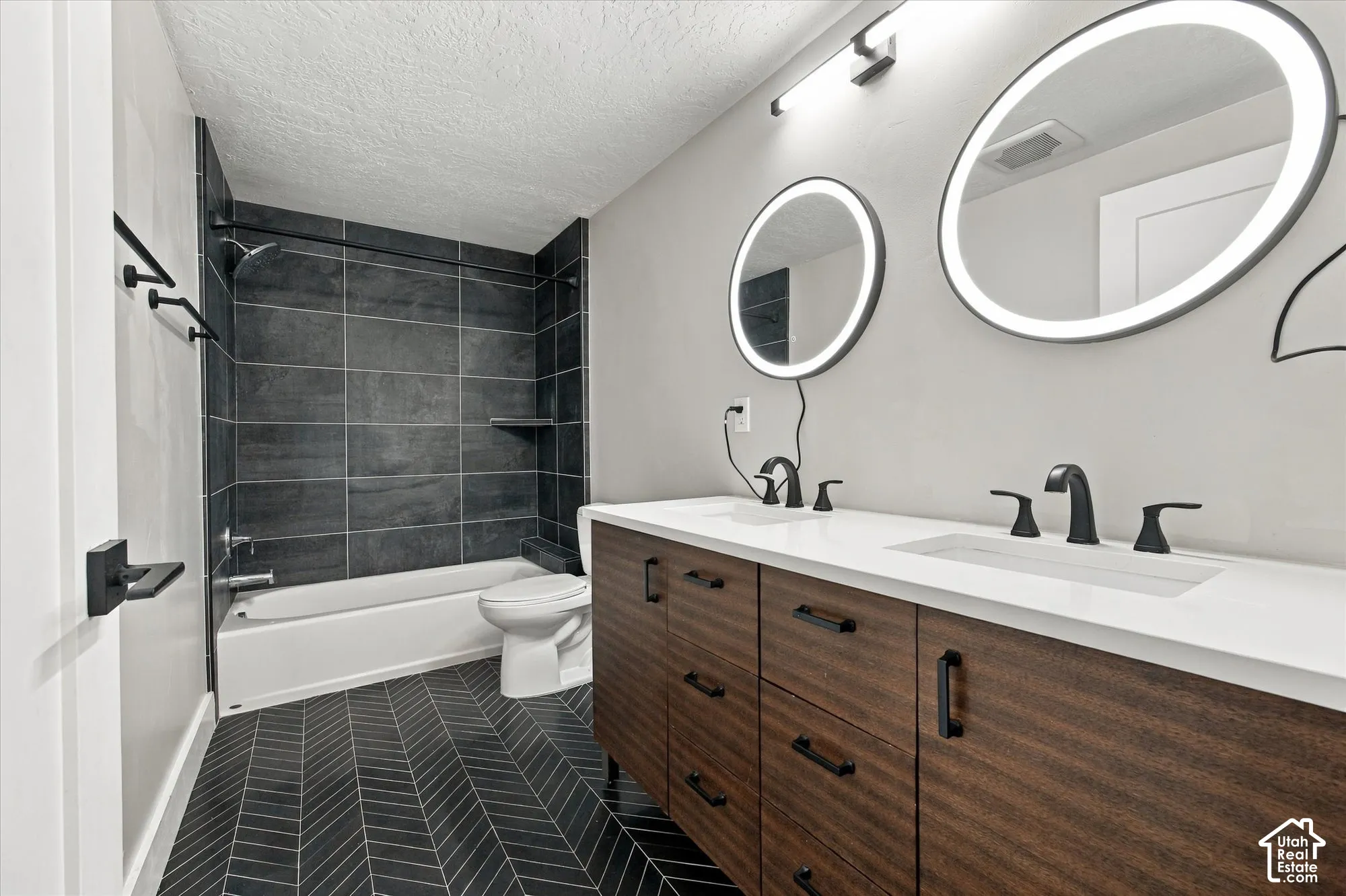 Double vanity featuring wood-finish cabinetry, white countertop, illuminated circular mirrors, matte black fixtures, and herringbone-patterned flooring