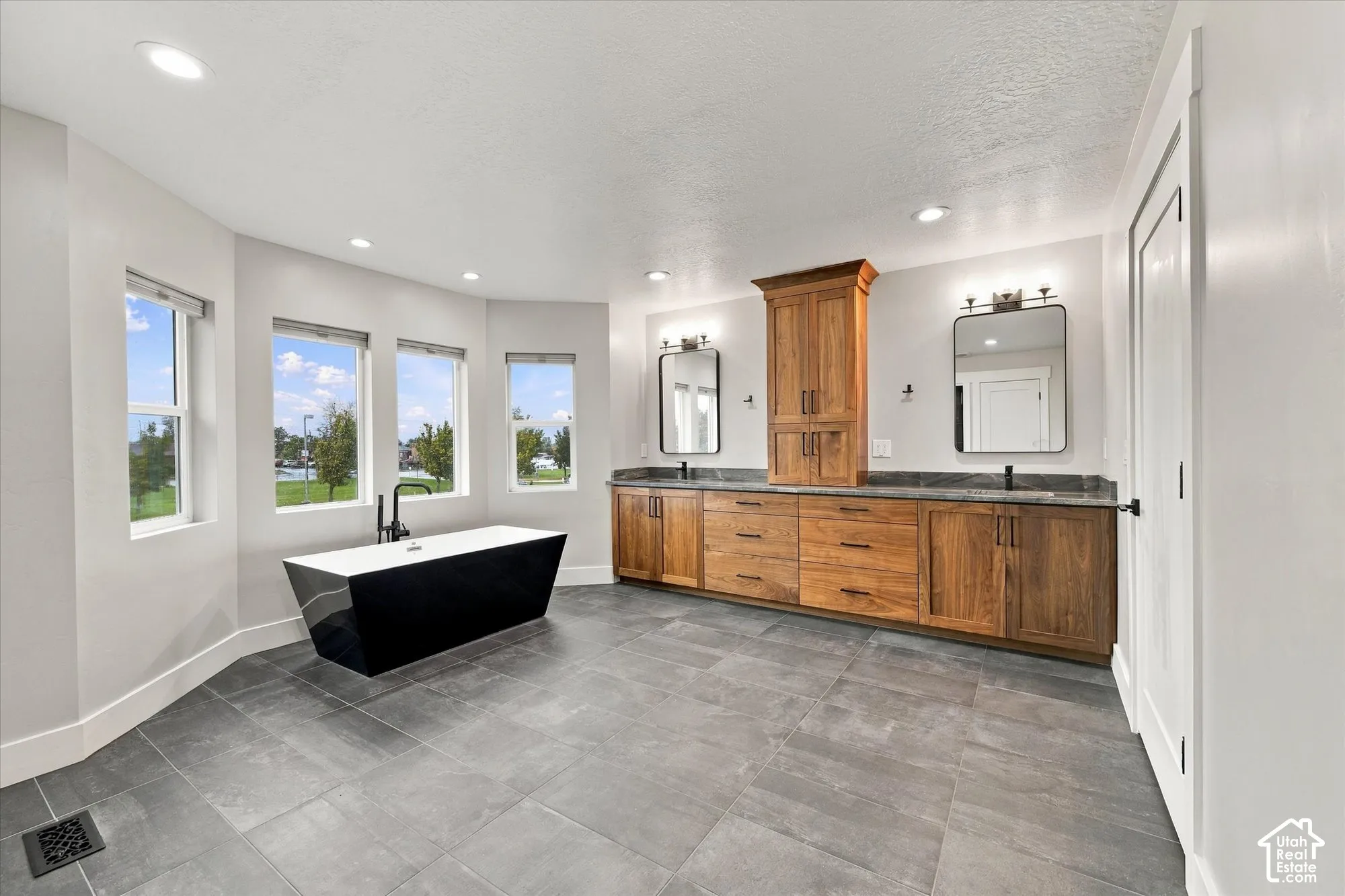 Freestanding soaking tub with a matte black faucet, custom wood-finish dual vanity with a tower cabinet, dark stone-style countertops, rectangular mirrors, and large-format tile flooring
