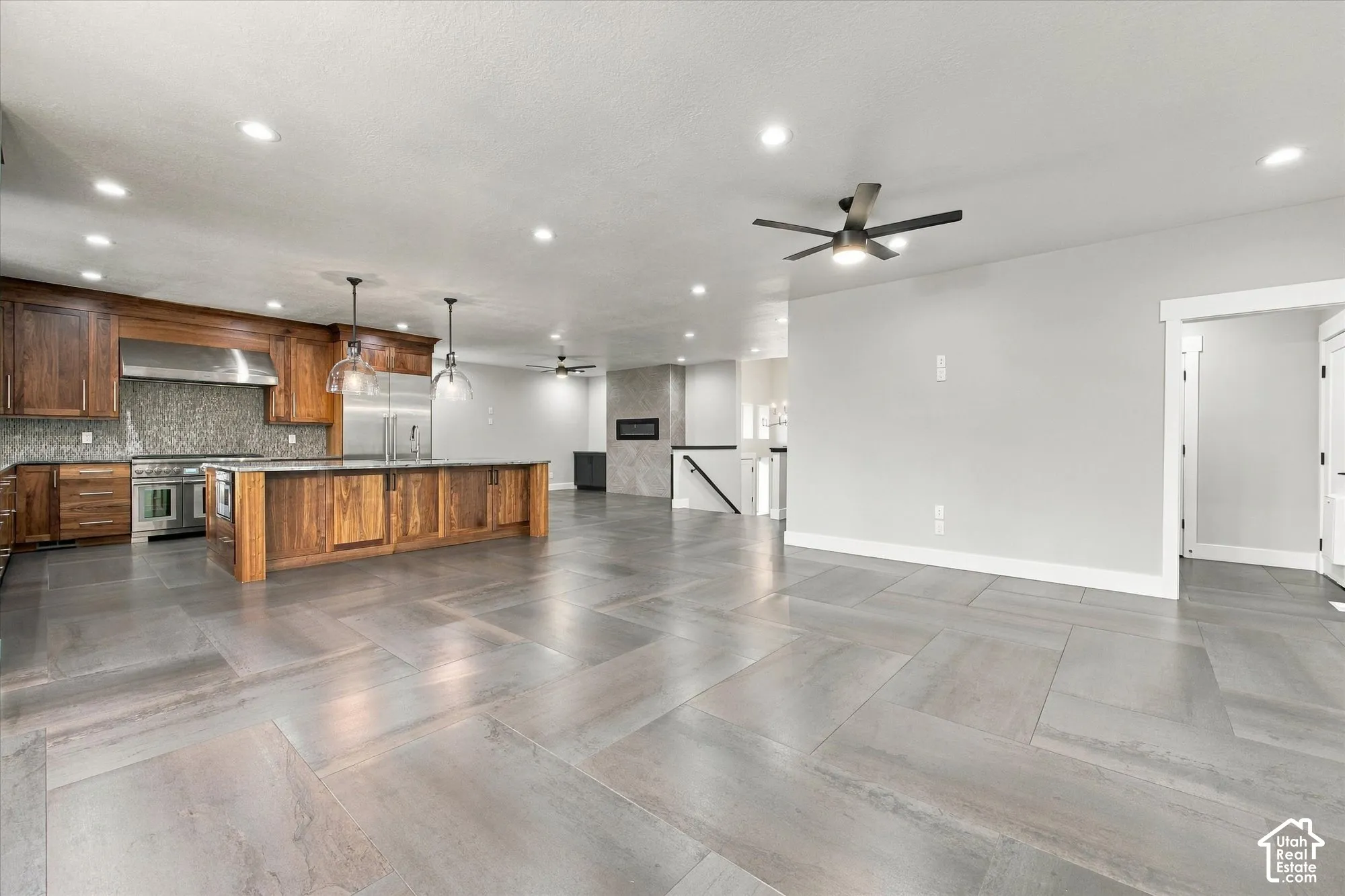 Open-concept interior featuring large-format tile flooring, wood-finish cabinetry, a stainless steel range with overhead hood, dual pendant lighting, and a built-in linear fireplace