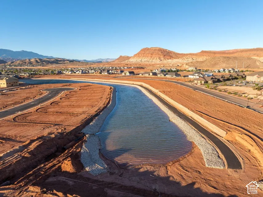 Aerial perspective of a development site featuring a central water basin, stone-lined perimeter, asphalt roadways, and mountain backdrop