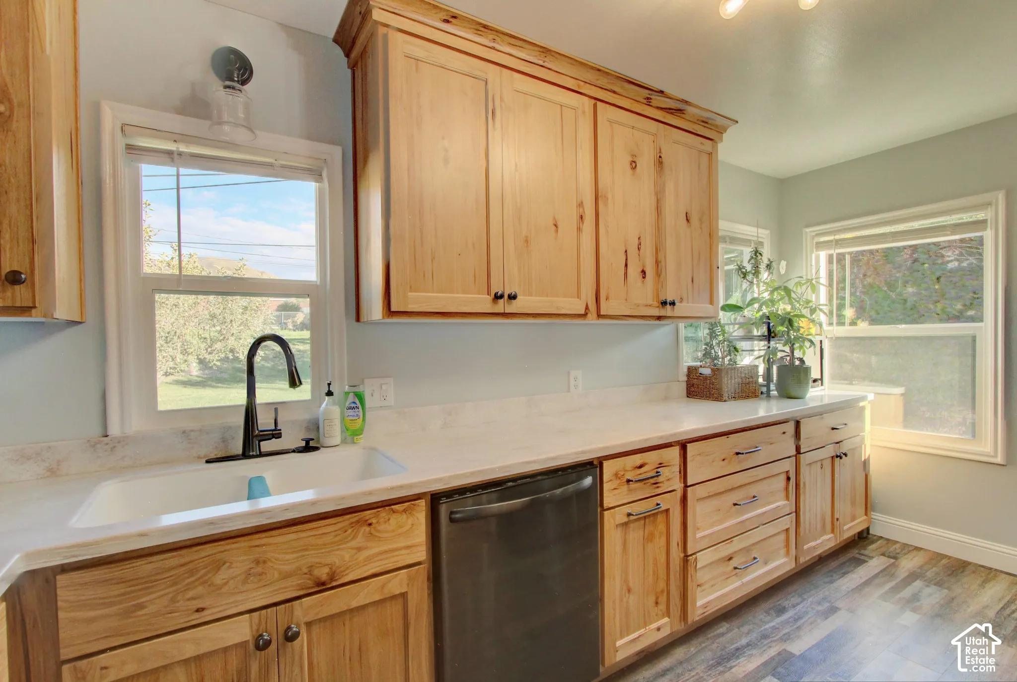 Newly remodeled. Natural wood-finish cabinetry, light-toned solid surface countertops, farmhouse-style sink with matte black faucet, integrated dishwasher, and wood-finish flooring