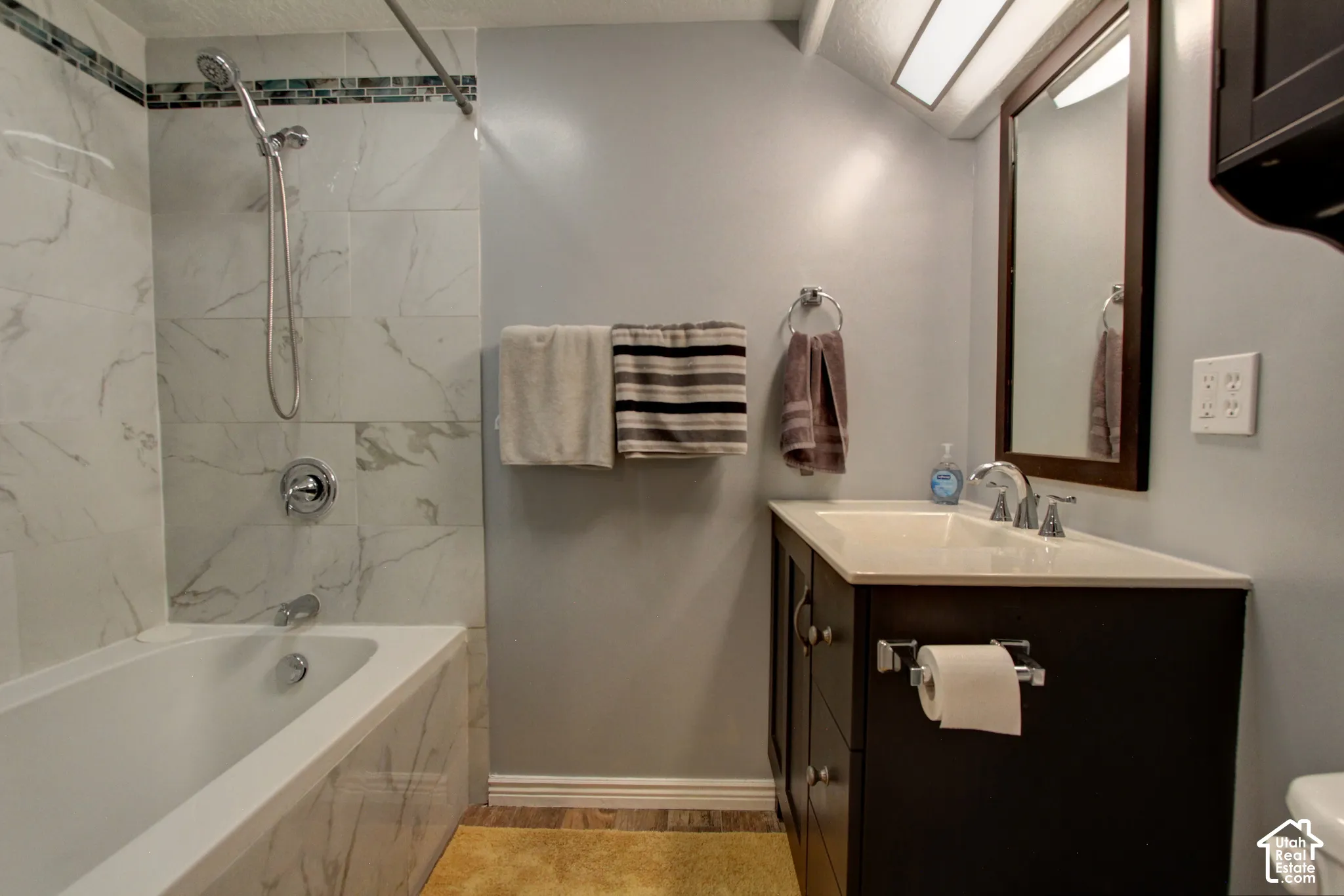 Full-wall tile surround with mosaic accent, deep soaking tub with handheld shower fixture, vanity with light-tone surface, and dark-finish built-in cabinetry