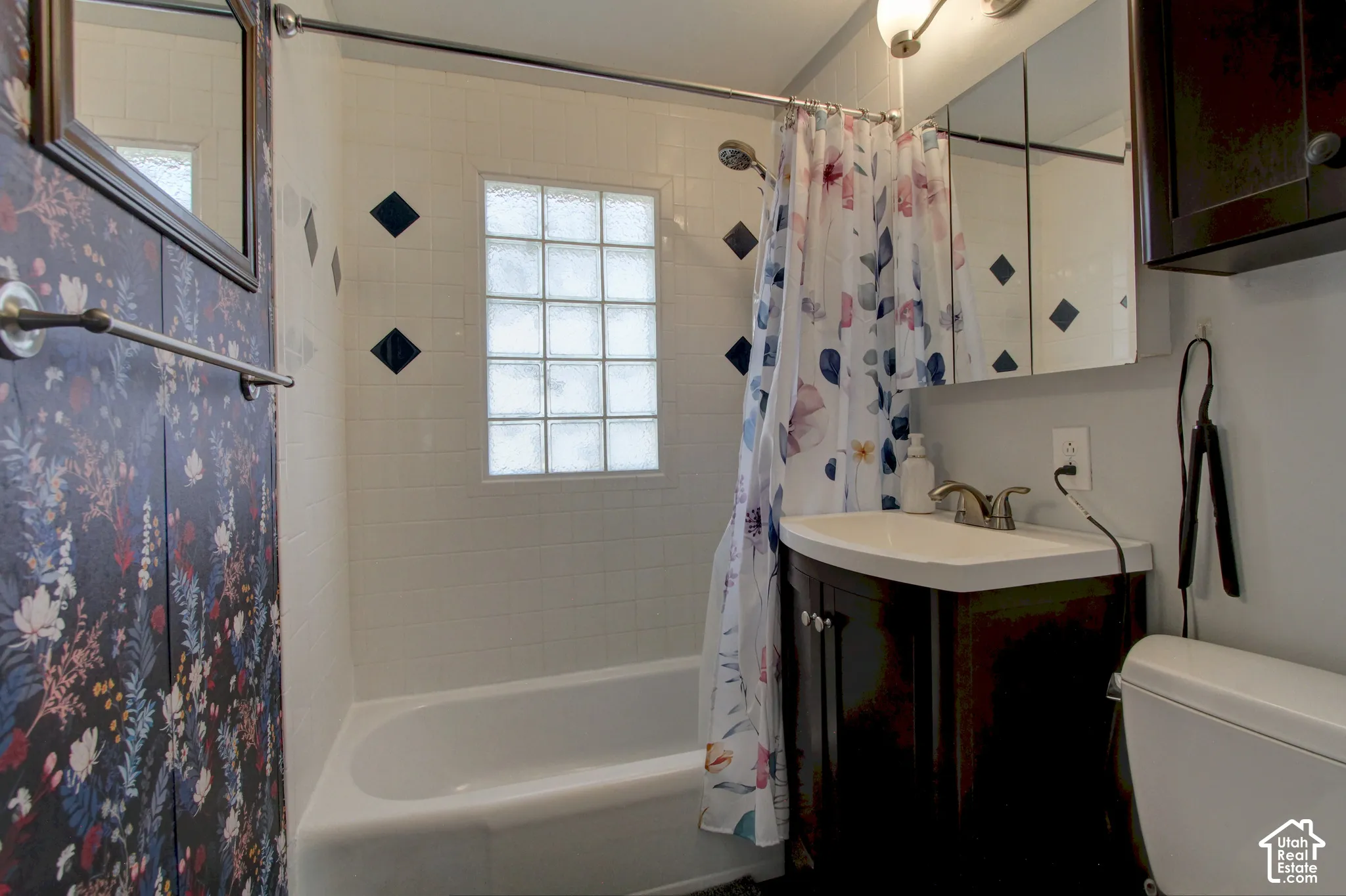 Bathroom featuring a vanity with a white basin and metallic faucet, glass block window, tile surround with decorative inlays, and a mirrored medicine cabinet