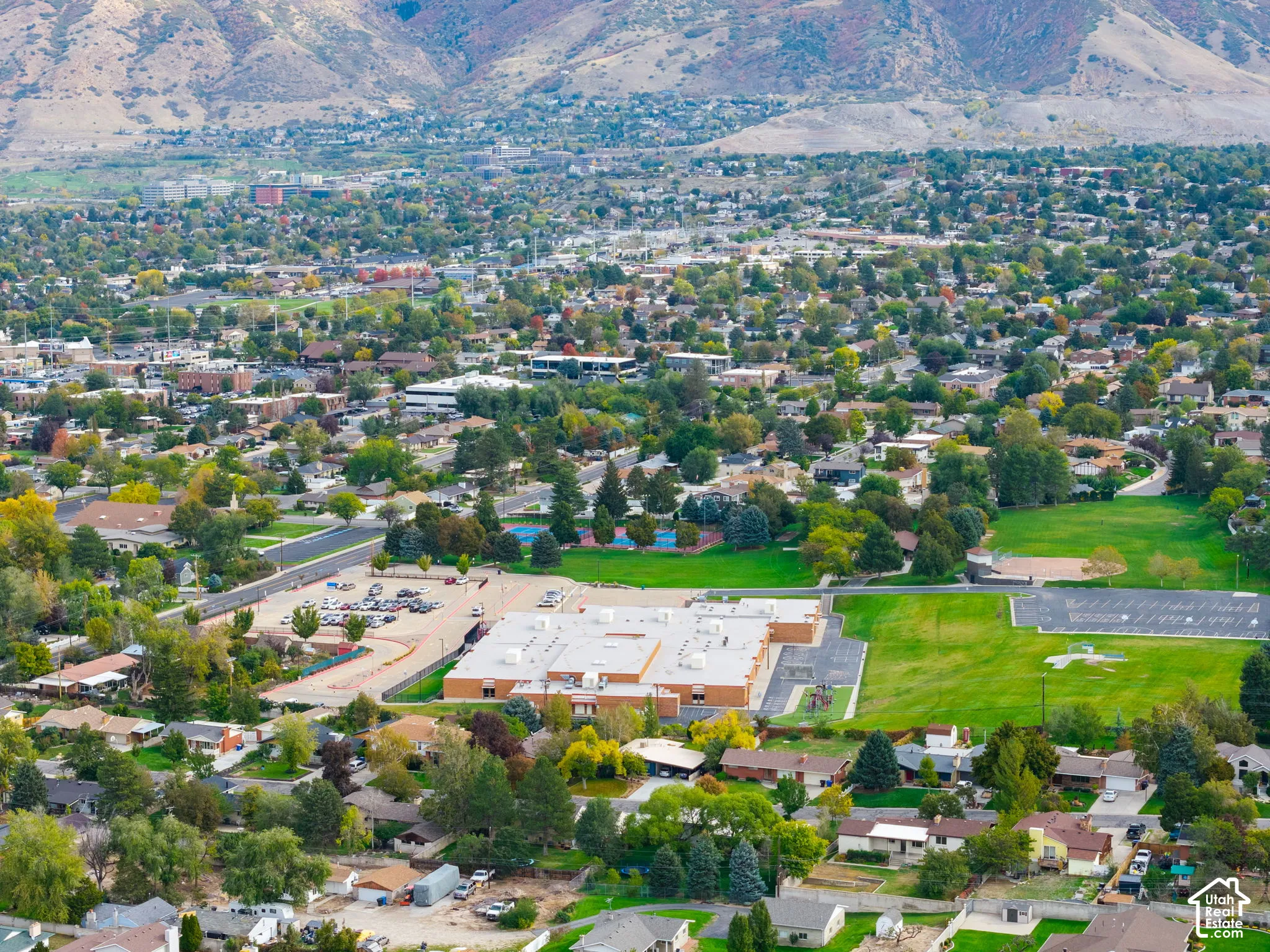 Aerial perspective highlighting a sprawling residential community set against a prominent mountain backdrop, featuring expansive green spaces, established tree canopy, and proximity to paved recreation facilities