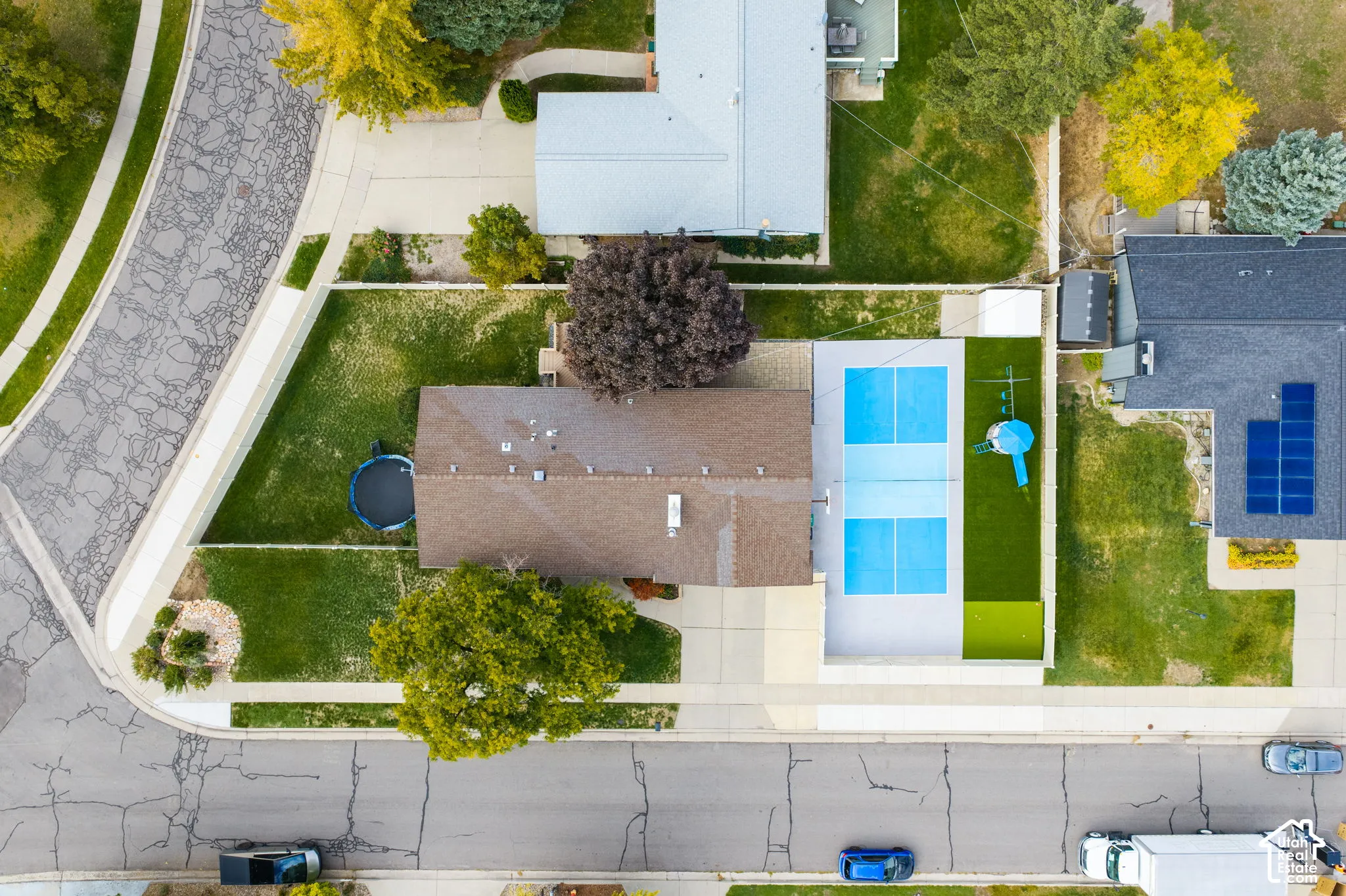 Aerial perspective of a corner lot property featuring a hardscaped court, fenced perimeter, mature trees, and an attached garage