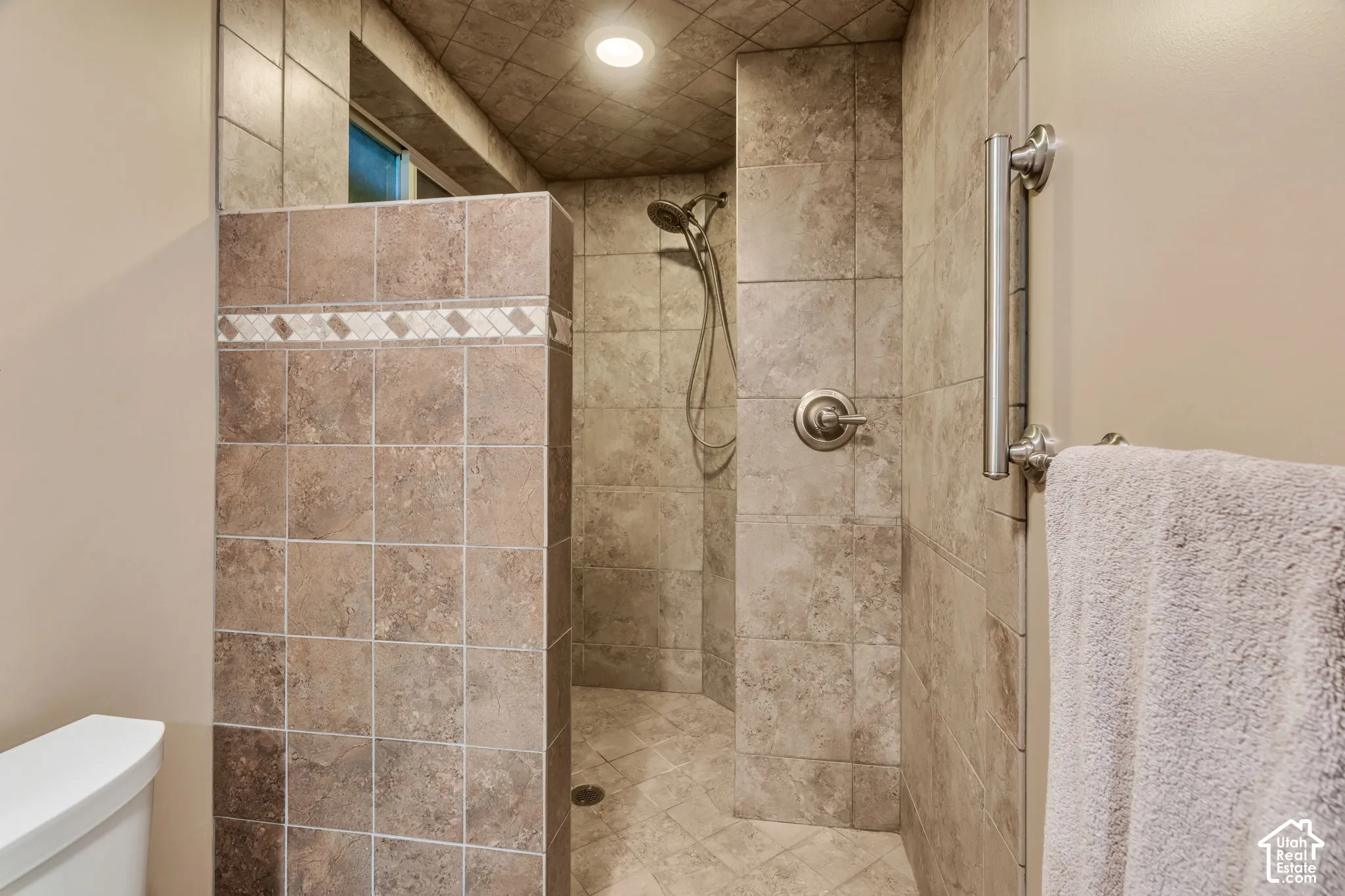 Walk-in shower featuring ceramic wall and floor tile, decorative tile inlay, a handheld showerhead, and a mounted stainless steel grab bar