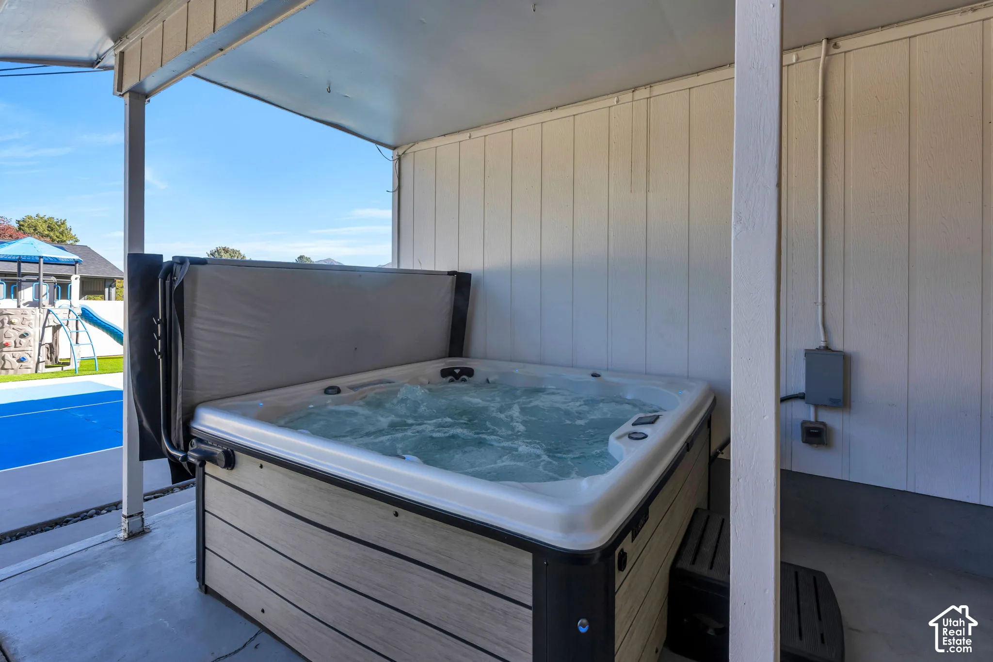 Covered outdoor patio area featuring a built-in hot tub with wood-finish siding, white vertical panel siding, and concrete flooring