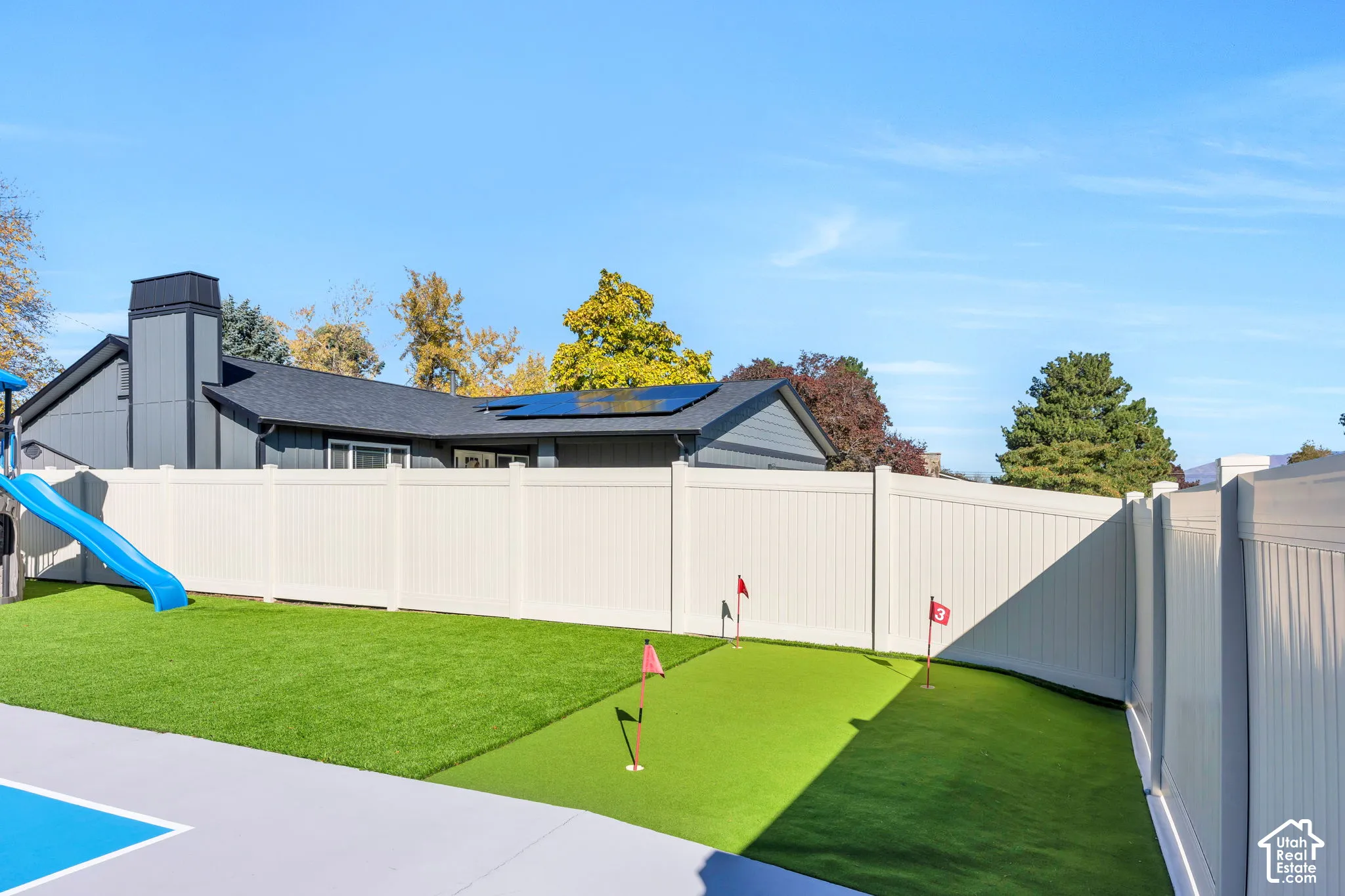 Fenced backyard featuring an artificial turf putting green with multiple holes, a concrete patio, and exterior solar panels