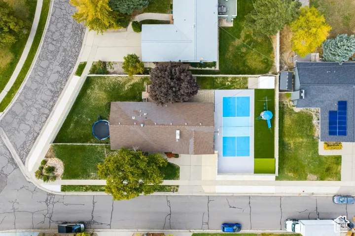 Aerial perspective of a corner lot property featuring a hardscaped court, fenced perimeter, mature trees, and an attached garage
