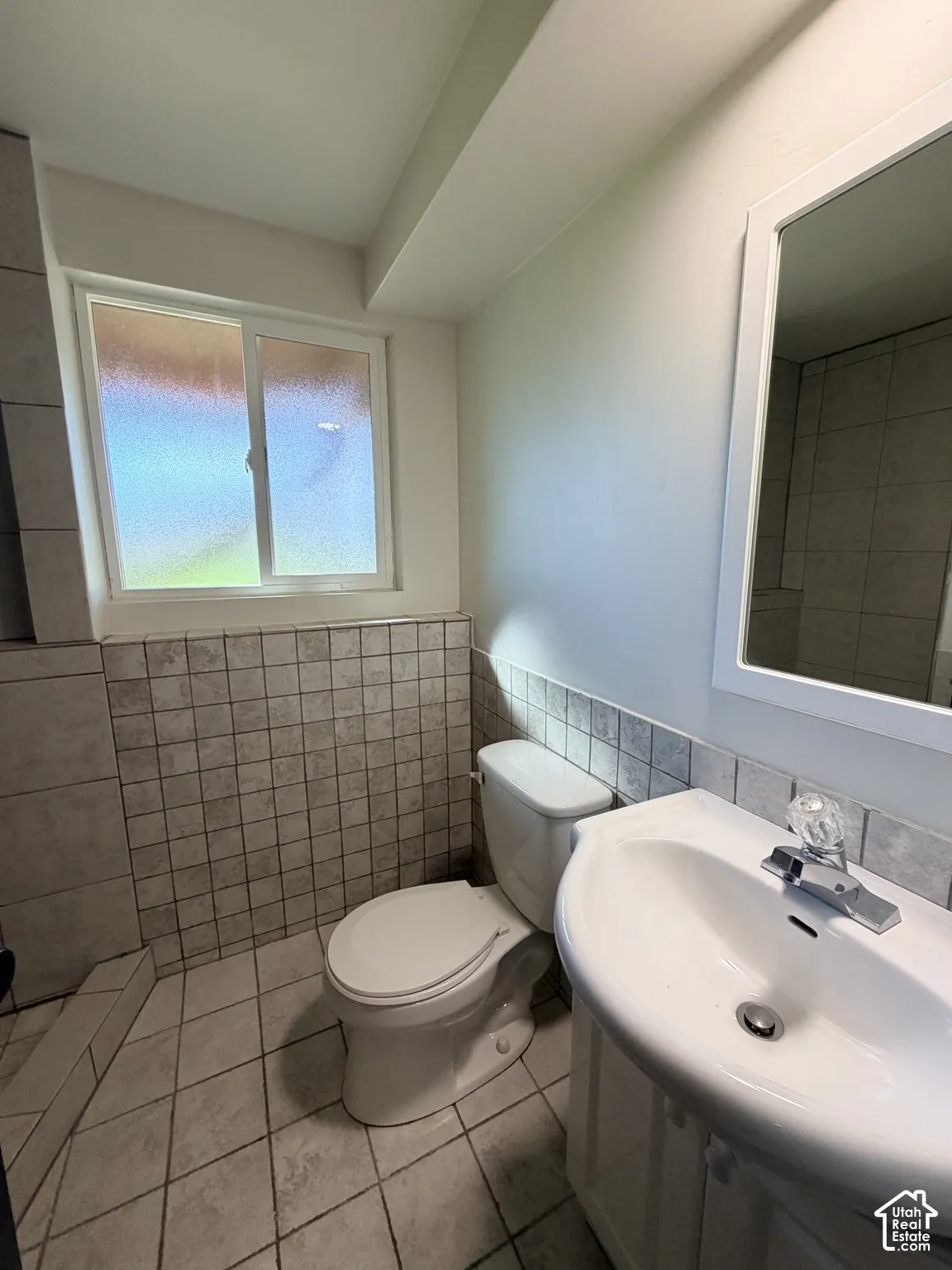 Bathroom featuring tile flooring, square wall tile wainscoting, sliding glass window, pedestal sink with chrome faucet, and a wall-mounted mirror with a white frame