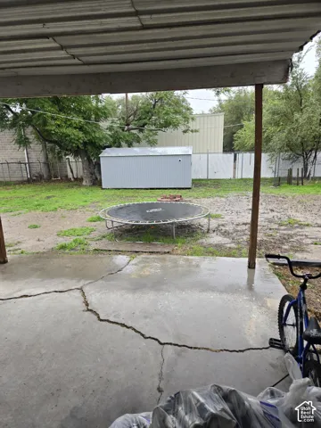 Covered concrete patio featuring an open-air backyard with perimeter fencing, mature trees, and a detached metal storage structure