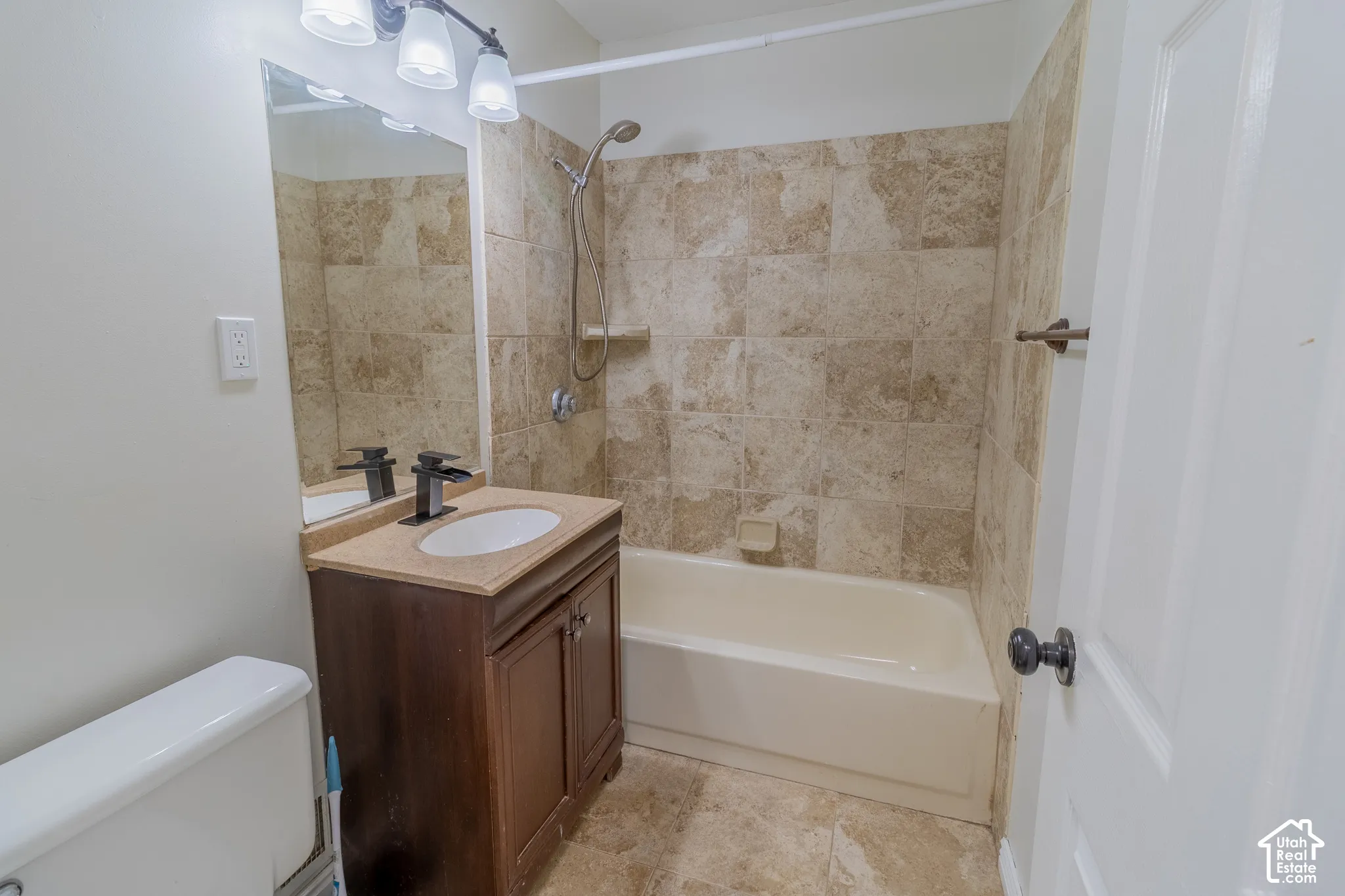Bathroom featuring a stone-tile tub surround, vanity with an integrated sink and stone countertop, three-light wall-mounted fixture, and shower head with handheld attachment