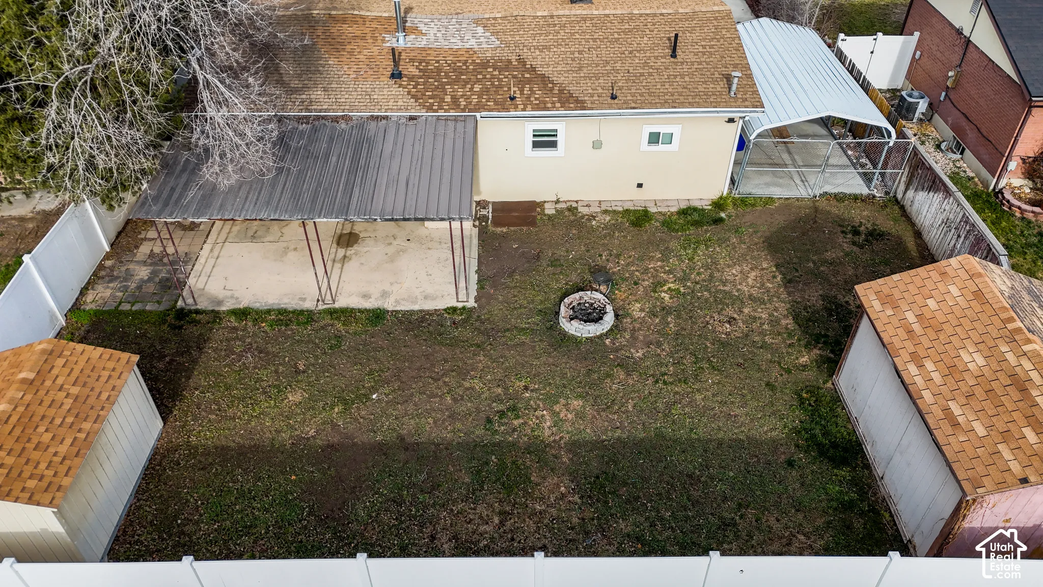 Fenced backyard featuring a covered patio, two detached storage structures, a stone-bordered fire pit, and a metal carport