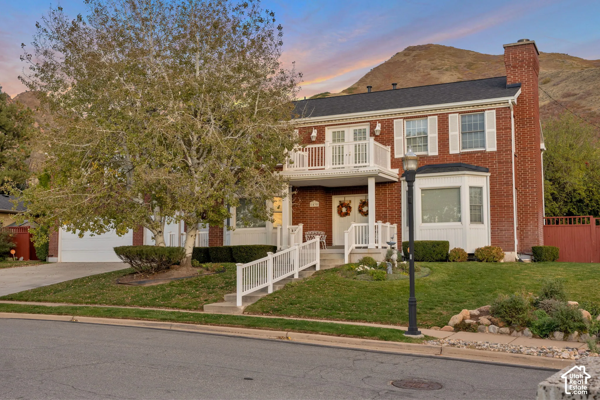 Two-story brick exterior featuring a covered front porch, second-story balcony with railings, external brick chimney, and a multi-paned bay window set against a mountain backdrop