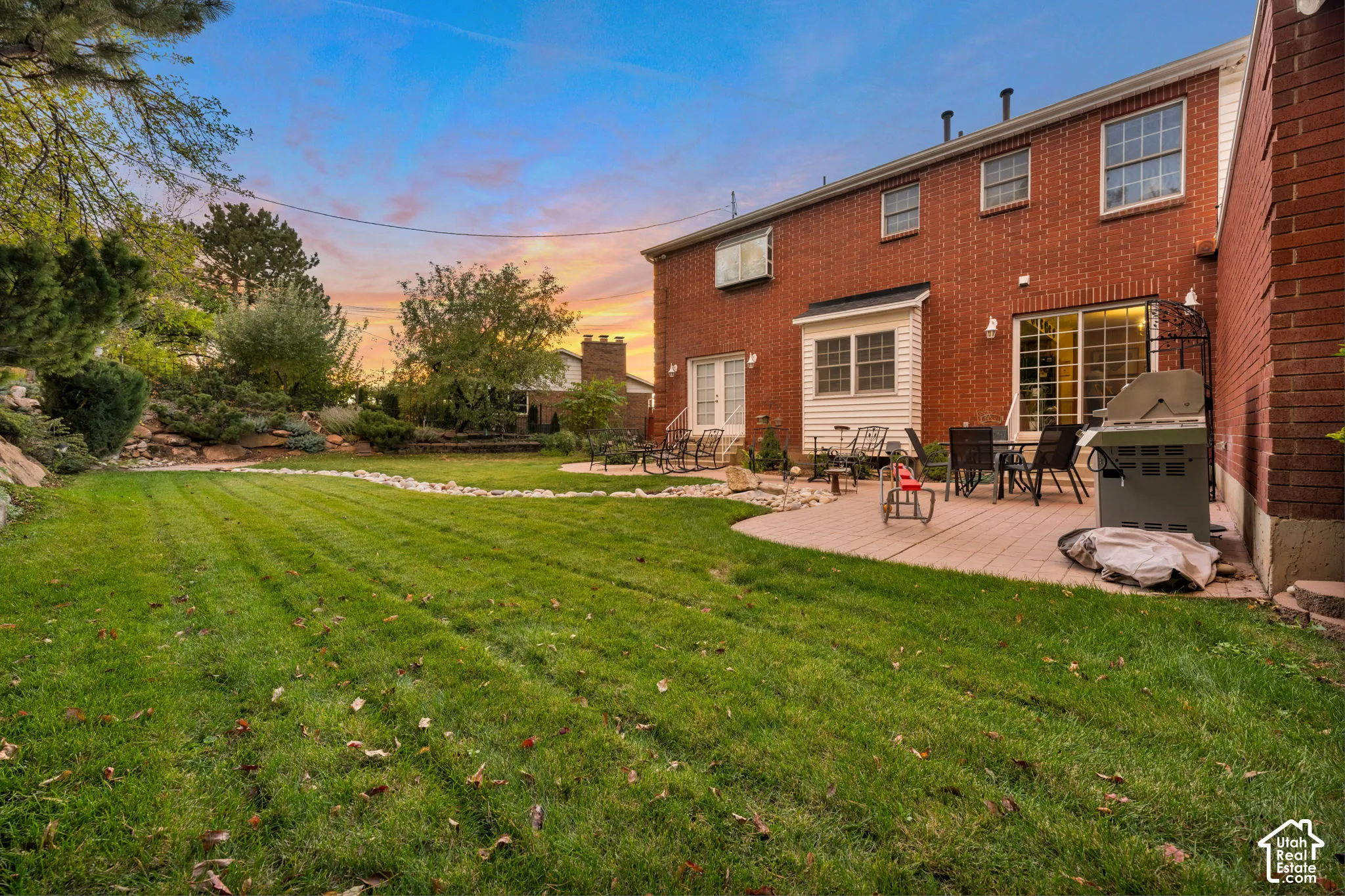 Red brick exterior featuring a multi-level paved patio, a manicured lawn, stone landscape edging, and mature trees