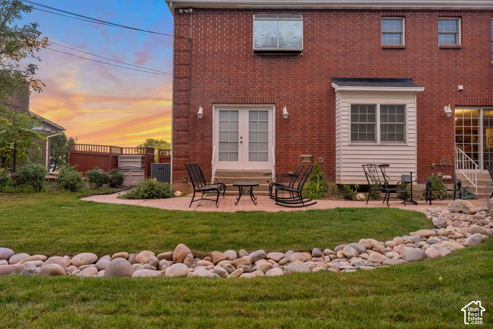 Brick exterior featuring a paved stone patio, dual glass-paned entry doors, a decorative river rock landscape border, and a manicured lawn