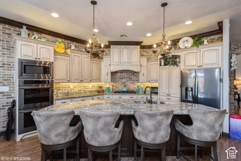 Kitchen featuring a large stone-surface island, cream-toned cabinetry with dark decorative molding, exposed brick walls, built-in stainless steel double ovens, and suspended decorative light fixtures