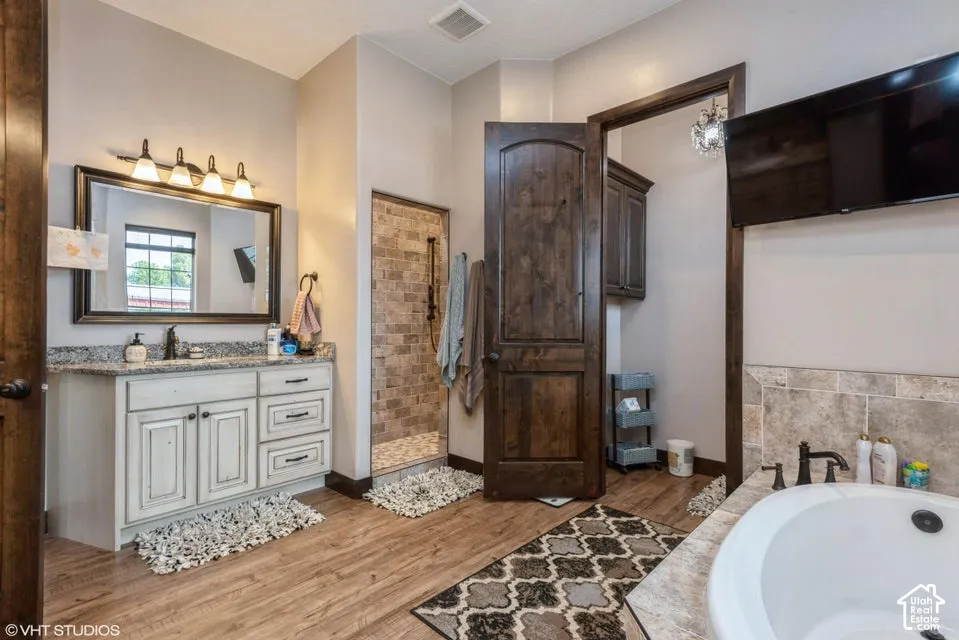 Bathroom featuring a custom vanity with stone countertop, tile-surround walk-in shower, soaking tub with stone deck, wood-finish cabinetry and doors, and wood-finish flooring