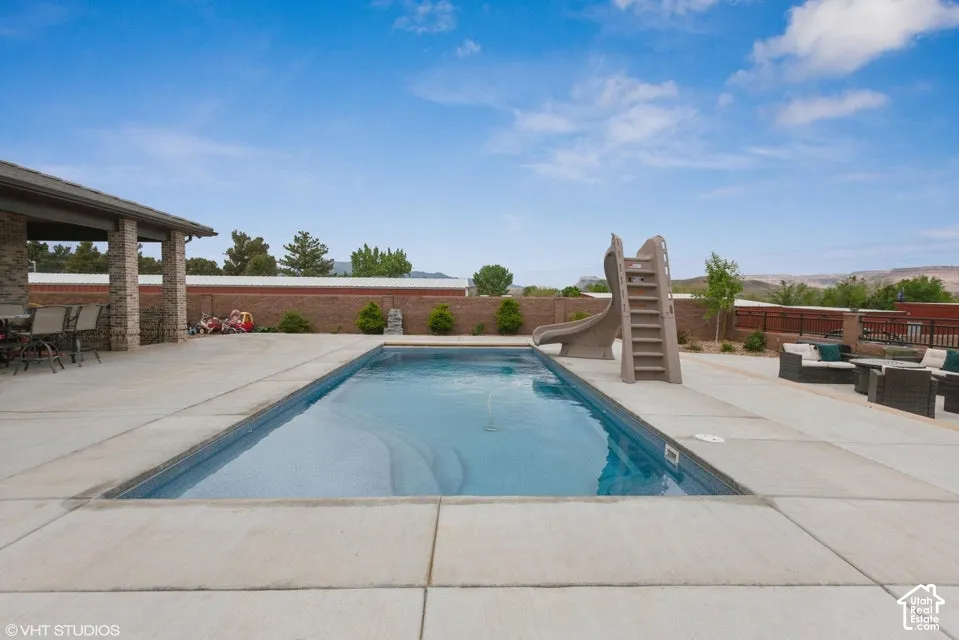 In-ground swimming pool with built-in slide, extensive concrete patio, covered outdoor area with stone pillars, and masonry perimeter wall