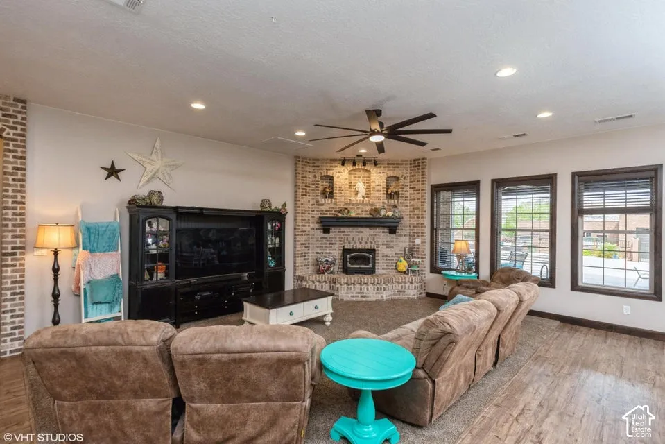 Corner fireplace with brick surround, wood-finish flooring, recessed lighting, ceiling fan, and large windows with dark trim