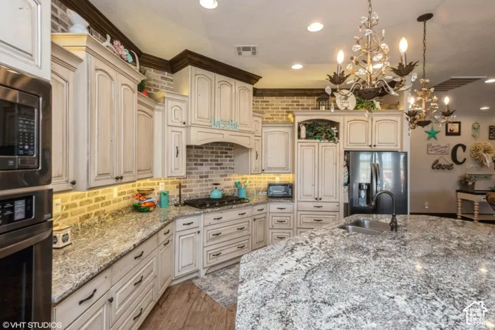 Kitchen featuring custom glazed cabinetry, stone countertops, exposed brick backsplash, wood-finish flooring, and dual decorative chandeliers